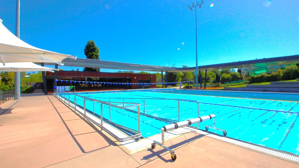 An outdoor swimming pool with clear blue water is surrounded by a paved area. On the left, there is a white canopy providing shade. A metal railing runs along the pool's edge. In the background, there are trees and a low building. A tall light pole is visible on the right. The sky is clear and blue. An outdoor swimming pool with clear blue water is surrounded by a paved area. On the left, there is a white canopy providing shade. A metal railing runs along the pool's edge. In the background, there are trees and a low building. A tall light pole is visible on the right. The sky is clear and blue.