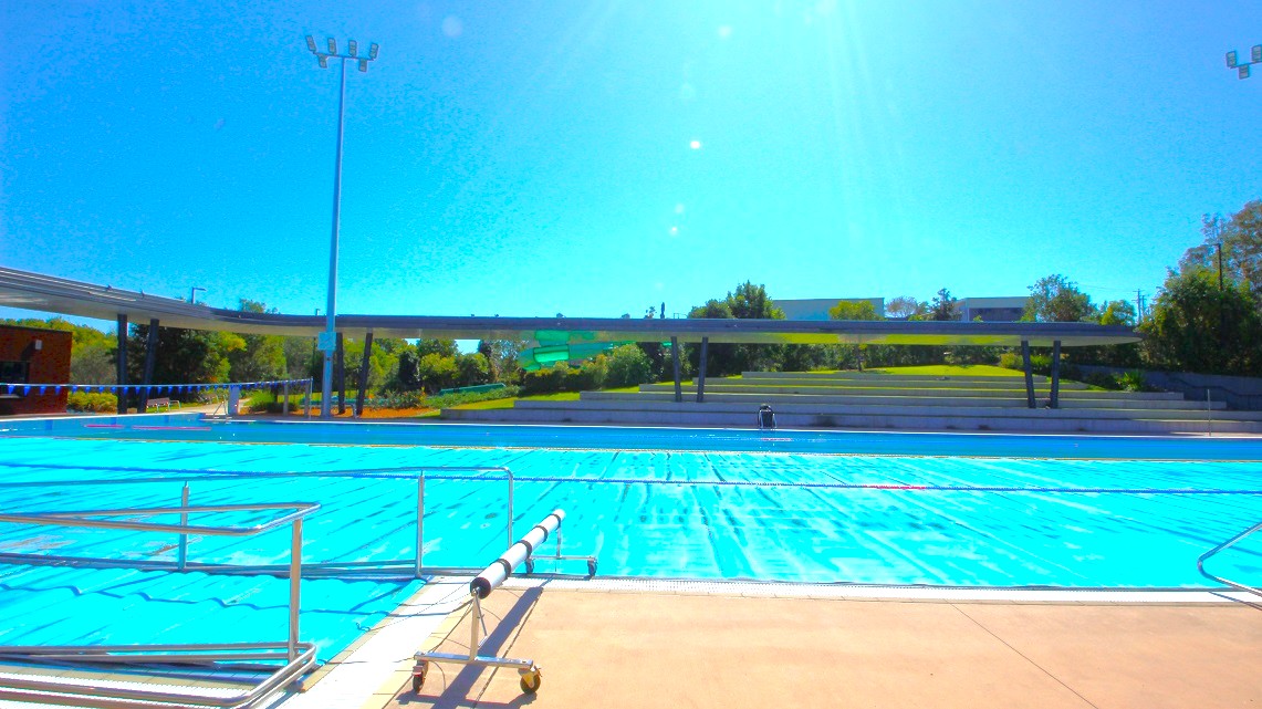 An outdoor swimming pool with clear blue water is in the foreground, surrounded by a beige pool deck. A metal railing runs along the pool's edge. In the background, there is a grassy area with trees and a few structures, including a long, low building. A tall light pole stands near the pool. The sky is clear and bright. An outdoor swimming pool with clear blue water is in the foreground, surrounded by a beige pool deck. A metal railing runs along the pool's edge. In the background, there is a grassy area with trees and a few structures, including a long, low building. A tall light pole stands near the pool. The sky is clear and bright.