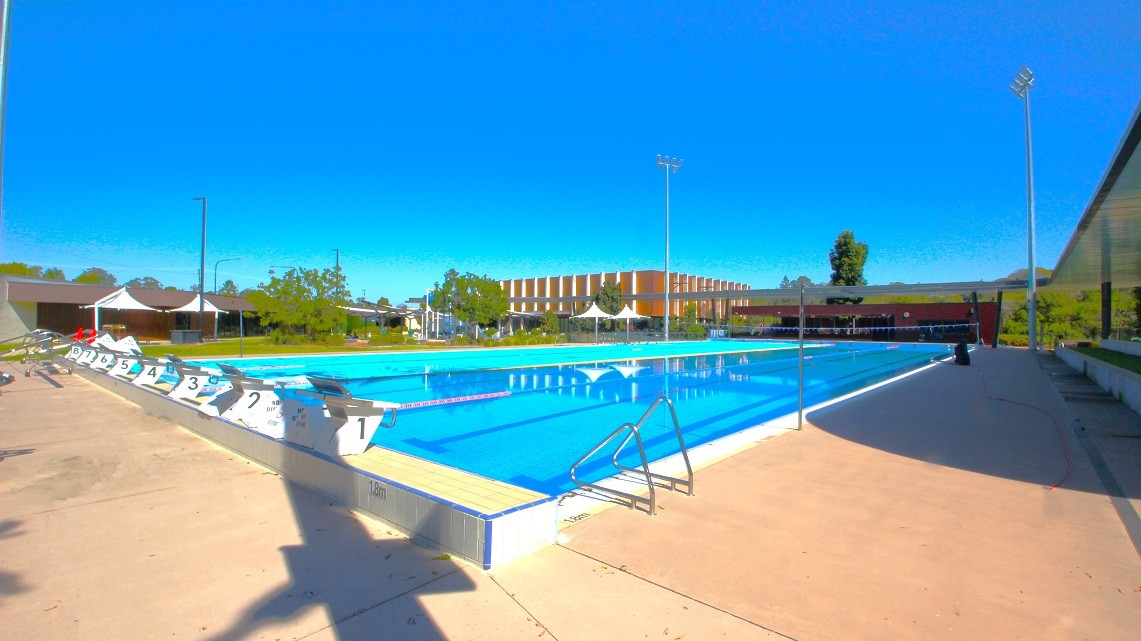 An outdoor swimming pool with clear blue water is surrounded by a concrete deck. On the left, there are numbered starting blocks and a row of white tents. In the background, a large building with a striped facade is visible, along with trees and a few streetlights. The right side features a shaded area with a railing. The sky is clear and blue, and the area is well-lit by tall floodlights. An outdoor swimming pool with clear blue water is surrounded by a concrete deck. On the left, there are numbered starting blocks and a row of white tents. In the background, a large building with a striped facade is visible, along with trees and a few streetlights. The right side features a shaded area with a railing. The sky is clear and blue, and the area is well-lit by tall floodlights.