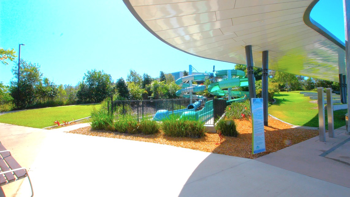 A water park scene with a large, twisting green water slide in the centre, surrounded by a black metal fence and lush greenery. The foreground has a paved walkway and a covered area with a curved roof supported by black pillars. On the left, there's a grassy area with trees in the background. The right side features more grass and a few small structures. The sky is clear and blue. A water park scene with a large, twisting green water slide in the centre, surrounded by a black metal fence and lush greenery. The foreground has a paved walkway and a covered area with a curved roof supported by black pillars. On the left, there's a grassy area with trees in the background. The right side features more grass and a few small structures. The sky is clear and blue.
