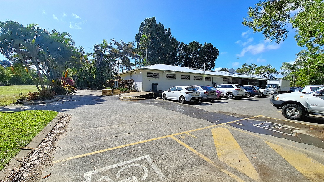 On the left, a white pickup truck is parked in a designated parking space, with a faded symbol indicating accessibility. The ground is a mix of asphalt and dirt, showing signs of wear. In the centre, yellow lines mark the parking area, leading towards a grassy field. On the right, a second accessible parking space is visible, also marked with a symbol. In the background, tall trees provide shade, and a patch of green grass extends further back. On the left, a white pickup truck is parked in a designated parking space, with a faded symbol indicating accessibility. The ground is a mix of asphalt and dirt, showing signs of wear. In the centre, yellow lines mark the parking area, leading towards a grassy field. On the right, a second accessible parking space is visible, also marked with a symbol. In the background, tall trees provide shade, and a patch of green grass extends further back.