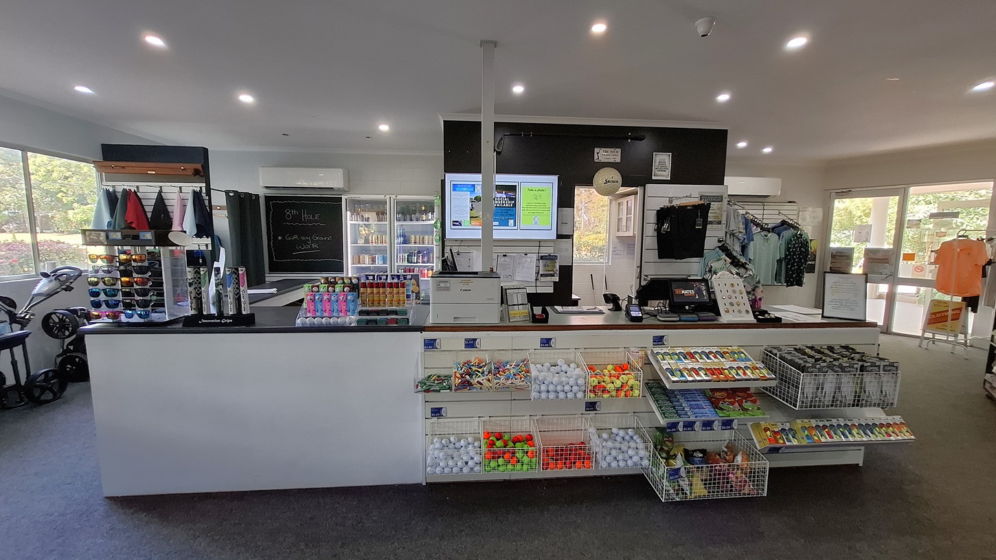 On the left, a display rack holds various coloured caps and sunglasses, with a golf trolley positioned nearby. The floor is covered in dark carpet, leading to a service counter in the centre. The counter features a cash register and a selection of snacks and golf balls in clear containers. On the right, a large glass door opens to the outside, with shirts hanging on a rack nearby. The ceiling is flat and white, with recessed lighting. On the left, a display rack holds various coloured caps and sunglasses, with a golf trolley positioned nearby. The floor is covered in dark carpet, leading to a service counter in the centre. The counter features a cash register and a selection of snacks and golf balls in clear containers. On the right, a large glass door opens to the outside, with shirts hanging on a rack nearby. The ceiling is flat and white, with recessed lighting.