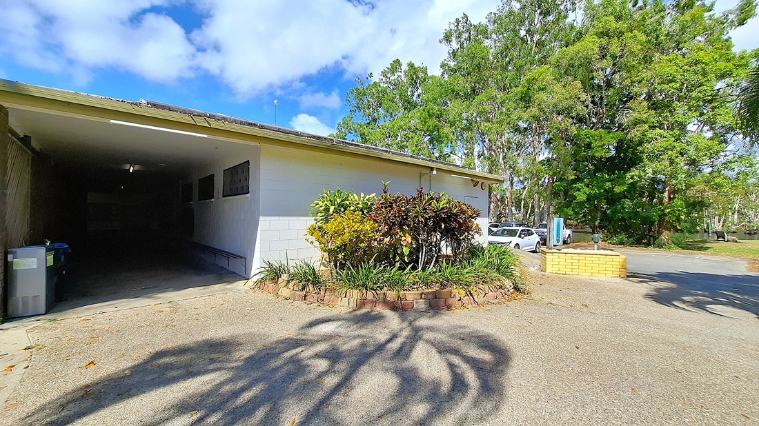 On the left, a garage entrance is visible, with a concrete floor and a few fluorescent lights illuminating the interior. The wall beside the garage is painted white, featuring several small windows. In the centre, a circular garden bed is surrounded by a low brick border, filled with various plants and shrubs. On the right, a white car is parked next to the garden, with a pathway leading further into the area. The ground is a mix of concrete and gravel. On the left, a garage entrance is visible, with a concrete floor and a few fluorescent lights illuminating the interior. The wall beside the garage is painted white, featuring several small windows. In the centre, a circular garden bed is surrounded by a low brick border, filled with various plants and shrubs. On the right, a white car is parked next to the garden, with a pathway leading further into the area. The ground is a mix of concrete and gravel.