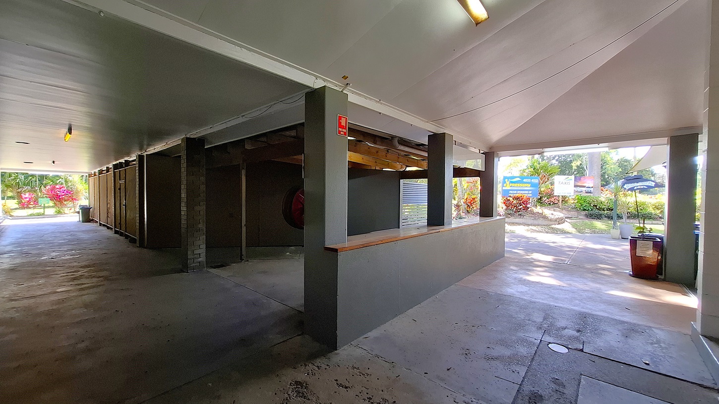 On the left, a concrete wall features a series of storage units, with a fire extinguisher mounted on the side. The floor is smooth and grey, extending towards an open area with a pathway leading outside. In the centre, a wooden counter runs along the wall, providing a surface for service or display. On the right, a large opening leads to a landscaped area with colourful plants and a sign visible in the background. The ceiling is flat and painted white, with fluorescent lights. On the left, a concrete wall features a series of storage units, with a fire extinguisher mounted on the side. The floor is smooth and grey, extending towards an open area with a pathway leading outside. In the centre, a wooden counter runs along the wall, providing a surface for service or display. On the right, a large opening leads to a landscaped area with colourful plants and a sign visible in the background. The ceiling is flat and painted white, with fluorescent lights.