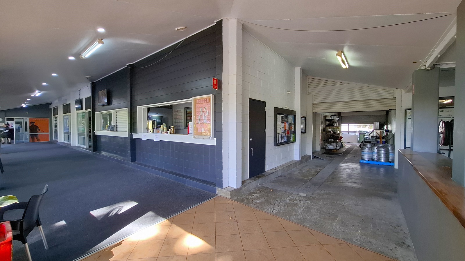 On the left, a long corridor features a dark carpeted floor, with several windows and a service counter displaying drinks and snacks. The walls are painted in light colours, with a few televisions mounted above. In the centre, the corridor leads to an open area with a tiled floor, transitioning to a concrete surface. On the right, a large opening reveals a storage area with kegs and equipment. The ceiling is flat and painted white, with fluorescent lights. On the left, a long corridor features a dark carpeted floor, with several windows and a service counter displaying drinks and snacks. The walls are painted in light colours, with a few televisions mounted above. In the centre, the corridor leads to an open area with a tiled floor, transitioning to a concrete surface. On the right, a large opening reveals a storage area with kegs and equipment. The ceiling is flat and painted white, with fluorescent lights.