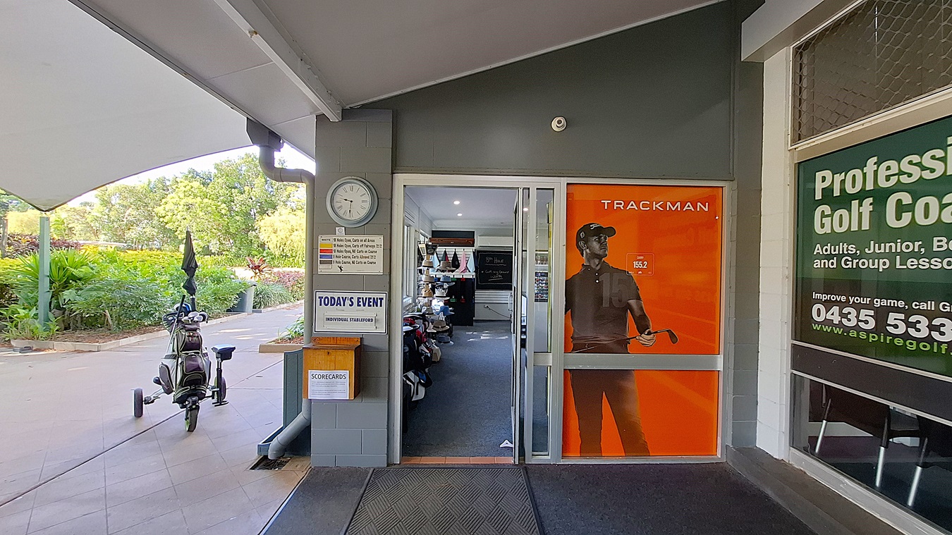 On the left, a clock is mounted on the wall beside a notice board displaying event information. A golf bag on wheels is parked nearby, with an umbrella attached. The entrance features a glass door with an orange advertisement for "TRACKMAN." In the centre, the floor is covered with dark carpet leading into the shop area, which has shelves filled with golf equipment. On the right, a large window displays information about golf coaching services. The ceiling is flat and white. On the left, a clock is mounted on the wall beside a notice board displaying event information. A golf bag on wheels is parked nearby, with an umbrella attached. The entrance features a glass door with an orange advertisement for "TRACKMAN." In the centre, the floor is covered with dark carpet leading into the shop area, which has shelves filled with golf equipment. On the right, a large window displays information about golf coaching services. The ceiling is flat and white.