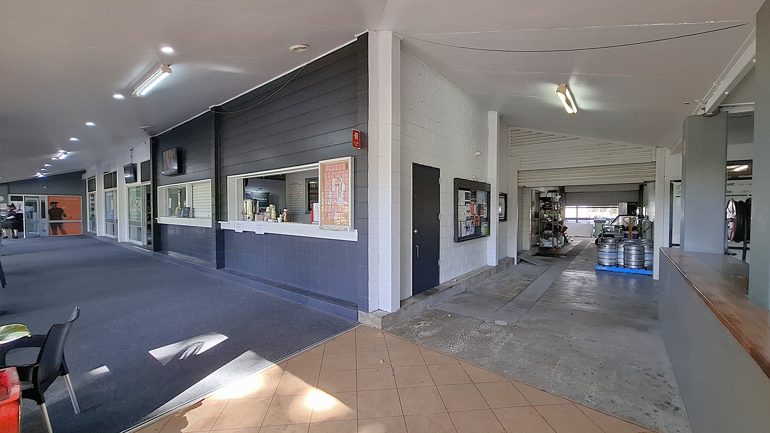 On the left, a long corridor features a dark carpeted floor, with several windows and a service counter displaying drinks and snacks. The walls are painted in light colours, with a few televisions mounted above. In the centre, the corridor leads to an open area with a tiled floor, transitioning to a concrete surface. On the right, a large opening reveals a storage area with kegs and equipment. The ceiling is flat and painted white, with fluorescent lights. On the left, a long corridor features a dark carpeted floor, with several windows and a service counter displaying drinks and snacks. The walls are painted in light colours, with a few televisions mounted above. In the centre, the corridor leads to an open area with a tiled floor, transitioning to a concrete surface. On the right, a large opening reveals a storage area with kegs and equipment. The ceiling is flat and painted white, with fluorescent lights.