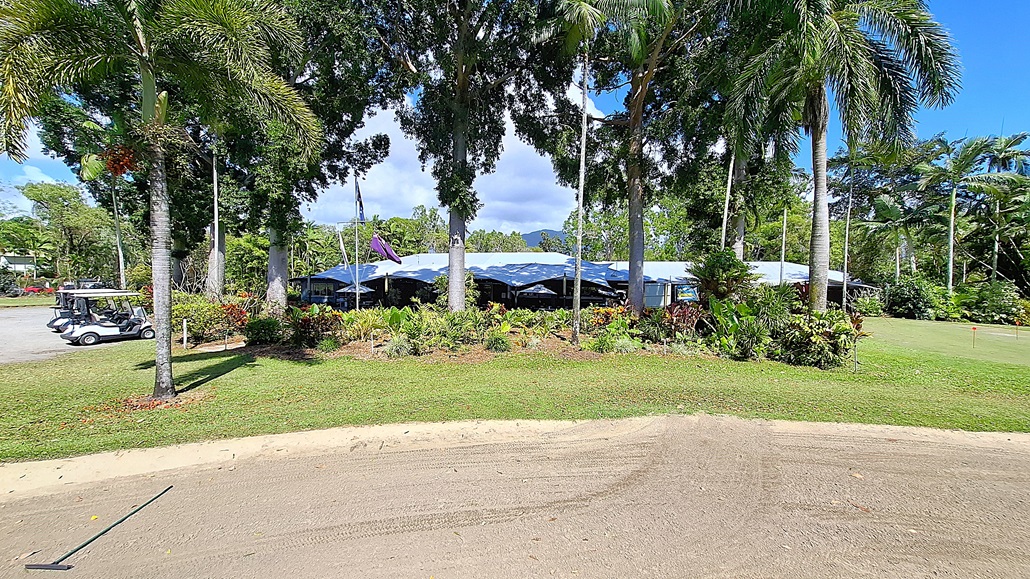 On the left, a row of golf carts is parked on a gravel surface, with a few palm trees providing shade. The ground is a mix of grass and sandy areas, leading towards a landscaped garden filled with various plants. In the centre, a building with a blue roof is partially obscured by trees, featuring large windows and outdoor seating areas. On the right, more greenery and trees create a natural border, enhancing the outdoor setting. On the left, a row of golf carts is parked on a gravel surface, with a few palm trees providing shade. The ground is a mix of grass and sandy areas, leading towards a landscaped garden filled with various plants. In the centre, a building with a blue roof is partially obscured by trees, featuring large windows and outdoor seating areas. On the right, more greenery and trees create a natural border, enhancing the outdoor setting.