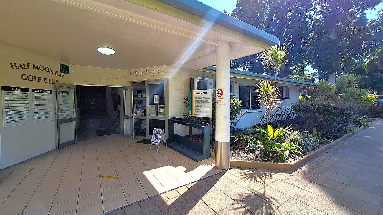 On the left, the entrance to the Half Moon Bay Golf Club features double glass doors, with a tiled floor leading inside. A notice board displays rules and etiquette for visitors. In the centre, the corridor extends into a darker area, while a service counter is visible to the right, with a sign detailing the dress code. On the right, a landscaped area includes various plants and a bench. The ceiling is flat and white, with a circular light fixture. On the left, the entrance to the Half Moon Bay Golf Club features double glass doors, with a tiled floor leading inside. A notice board displays rules and etiquette for visitors. In the centre, the corridor extends into a darker area, while a service counter is visible to the right, with a sign detailing the dress code. On the right, a landscaped area includes various plants and a bench. The ceiling is flat and white, with a circular light fixture.
