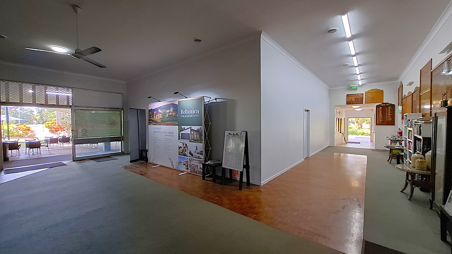 On the left, a large display stand features promotional materials for a local business, with a small table beside it. The floor is a mix of light-coloured tiles and a darker wooden section. In the centre, the corridor leads towards a doorway, with a light grey carpet underfoot. On the right, a wall showcases wooden plaques and a small shelf with items for sale. The ceiling is flat and white, with fluorescent lights. On the left, a large display stand features promotional materials for a local business, with a small table beside it. The floor is a mix of light-coloured tiles and a darker wooden section. In the centre, the corridor leads towards a doorway, with a light grey carpet underfoot. On the right, a wall showcases wooden plaques and a small shelf with items for sale. The ceiling is flat and white, with fluorescent lights.