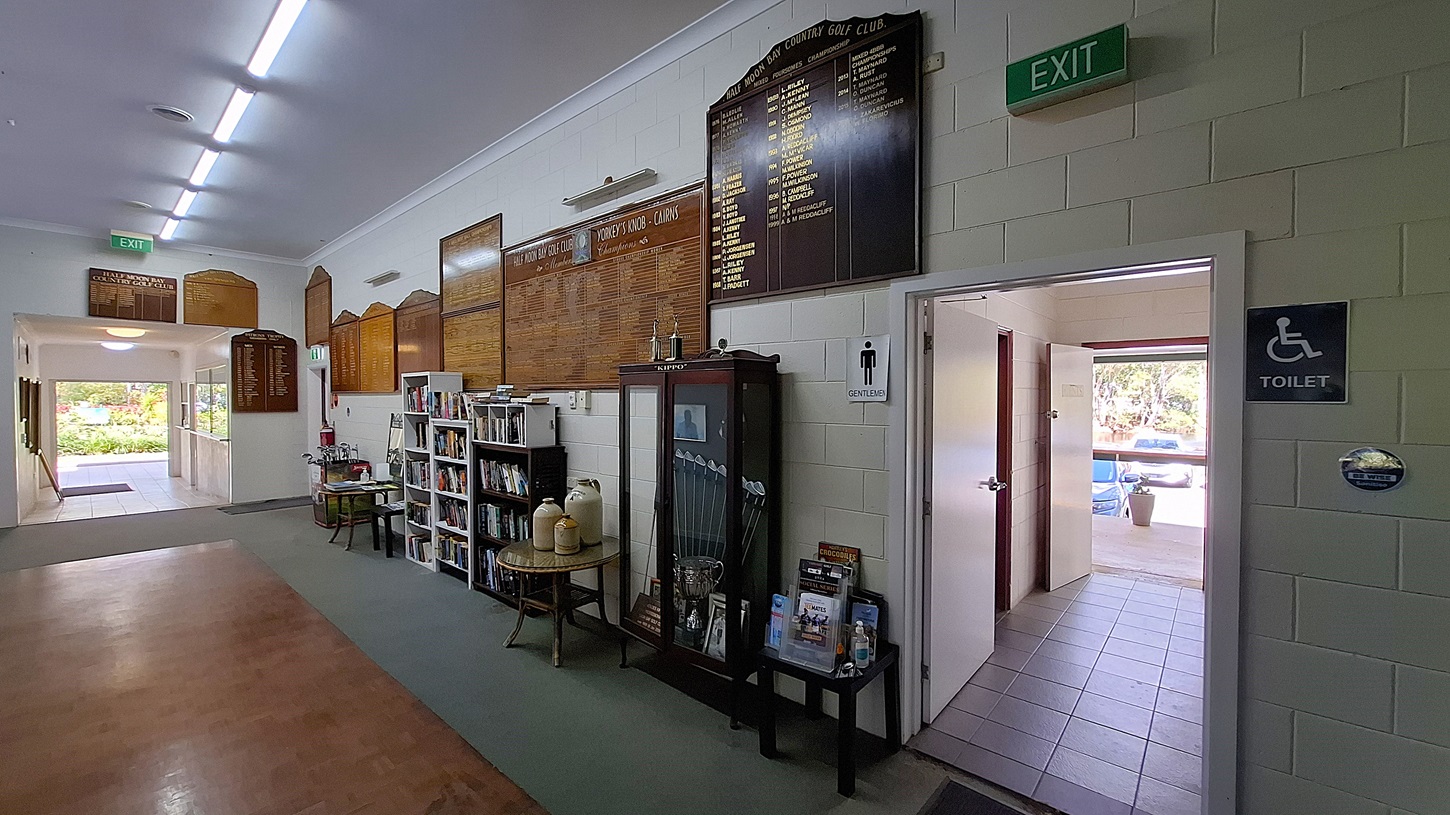 On the left, a bookshelf holds various books and pamphlets, with a small table displaying two large containers. The floor is a combination of light tiles and a darker carpet. In the centre, a corridor leads to an exit, with large wooden boards mounted on the wall displaying names and scores. On the right, a doorway leads to the toilets, marked with signs for "Gentlemen" and "Toilet." The ceiling is flat and white, with fluorescent lights. On the left, a bookshelf holds various books and pamphlets, with a small table displaying two large containers. The floor is a combination of light tiles and a darker carpet. In the centre, a corridor leads to an exit, with large wooden boards mounted on the wall displaying names and scores. On the right, a doorway leads to the toilets, marked with signs for "Gentlemen" and "Toilet." The ceiling is flat and white, with fluorescent lights.