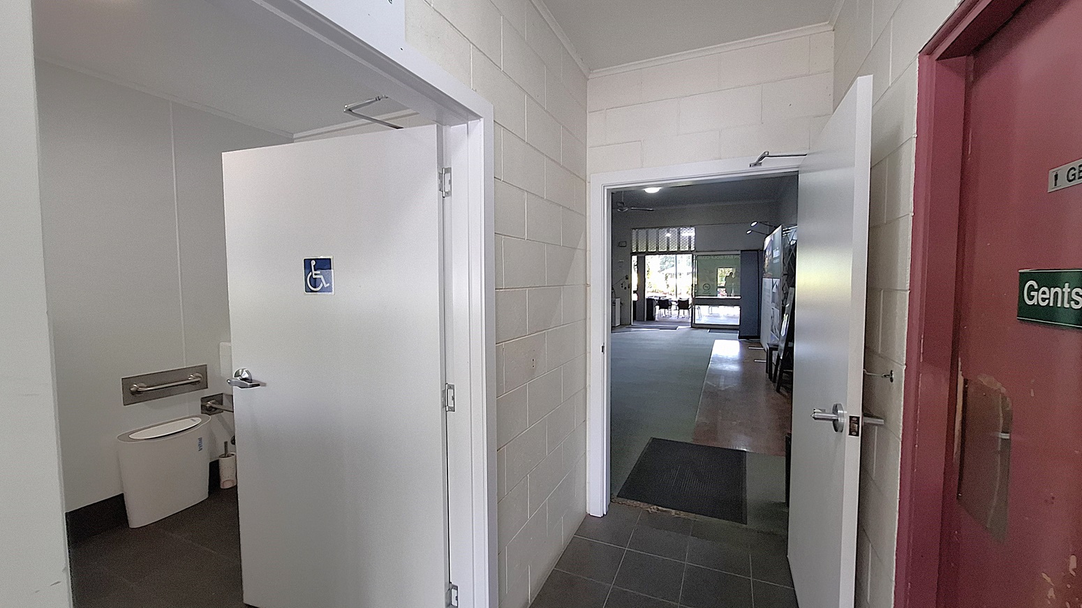 On the left, a white door leads to a disabled toilet, with a visible toilet and grab bars inside. The floor is dark tiled, contrasting with the light-coloured walls. In the centre, a corridor extends towards a larger room, with a smooth grey floor and a doorway leading outside. On the right, a red door is marked "Gents," indicating the men's toilet. The ceiling is flat and white, with overhead lights illuminating the area. On the left, a white door leads to a disabled toilet, with a visible toilet and grab bars inside. The floor is dark tiled, contrasting with the light-coloured walls. In the centre, a corridor extends towards a larger room, with a smooth grey floor and a doorway leading outside. On the right, a red door is marked "Gents," indicating the men's toilet. The ceiling is flat and white, with overhead lights illuminating the area.