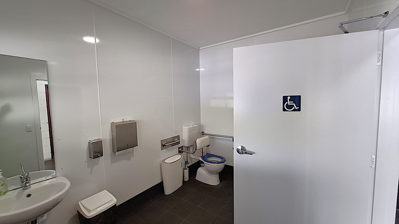 On the left, a white sink with a chrome tap is mounted on the wall, next to a small bin. Above the sink, a mirror reflects the space. The floor is dark tiled, providing a contrast to the light-coloured walls. In the centre, a toilet with a blue seat is positioned beside a wall-mounted grab rail. On the right, a white door marked with a blue accessibility symbol leads to a disabled toilet. The ceiling is flat and white, with overhead lights illuminating the area. On the left, a white sink with a chrome tap is mounted on the wall, next to a small bin. Above the sink, a mirror reflects the space. The floor is dark tiled, providing a contrast to the light-coloured walls. In the centre, a toilet with a blue seat is positioned beside a wall-mounted grab rail. On the right, a white door marked with a blue accessibility symbol leads to a disabled toilet. The ceiling is flat and white, with overhead lights illuminating the area.