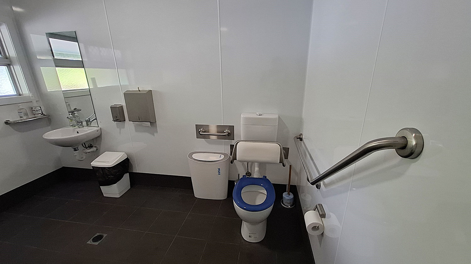 On the left, a white sink is mounted on the wall, with a mirror above it reflecting the space. A small shelf holds toiletries, and a bin is positioned below the sink. The floor is dark tiled, providing a contrast to the light walls. In the centre, a toilet with a blue seat is equipped with grab bars for accessibility. On the right, a roll of toilet paper is mounted on the wall, next to a silver handrail. The ceiling is flat and white, with overhead lights. On the left, a white sink is mounted on the wall, with a mirror above it reflecting the space. A small shelf holds toiletries, and a bin is positioned below the sink. The floor is dark tiled, providing a contrast to the light walls. In the centre, a toilet with a blue seat is equipped with grab bars for accessibility. On the right, a roll of toilet paper is mounted on the wall, next to a silver handrail. The ceiling is flat and white, with overhead lights.