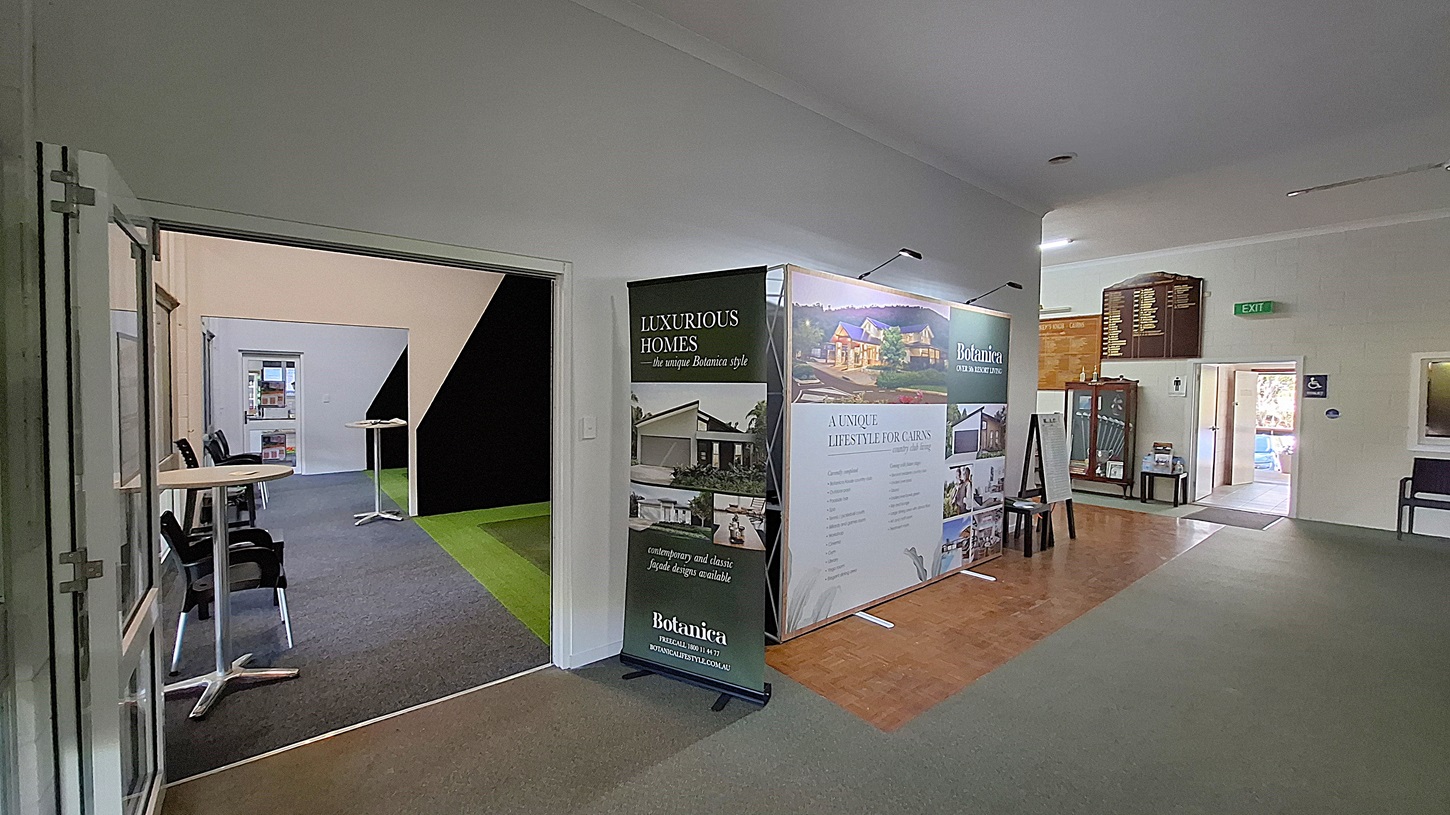 On the left, a large display stand features promotional materials for luxurious homes, with images and text highlighting various properties. A small table with two chairs is positioned nearby. The floor is a mix of dark carpet and wooden tiles. In the centre, a corridor leads to another room, with a green carpet visible on the right side. On the right, a wooden board lists names and achievements, and a doorway leads to a restroom. The ceiling is flat and white, with overhead lights. On the left, a large display stand features promotional materials for luxurious homes, with images and text highlighting various properties. A small table with two chairs is positioned nearby. The floor is a mix of dark carpet and wooden tiles. In the centre, a corridor leads to another room, with a green carpet visible on the right side. On the right, a wooden board lists names and achievements, and a doorway leads to a restroom. The ceiling is flat and white, with overhead lights.
