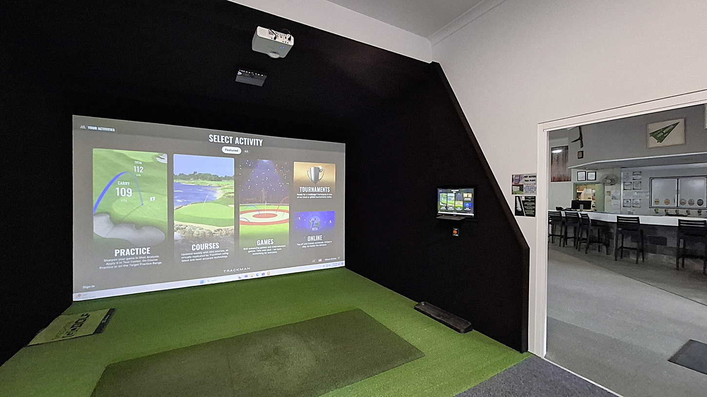 On the left, a projection screen displays options for golf activities, including practice and courses, with a green mat positioned in front for play. The floor is covered in artificial grass. In the centre, the space is enclosed by black walls, creating a focused environment. On the right, a small monitor shows additional information, and a doorway leads to another room with tables and chairs visible. The ceiling is flat and white, with a projector mounted above. On the left, a projection screen displays options for golf activities, including practice and courses, with a green mat positioned in front for play. The floor is covered in artificial grass. In the centre, the space is enclosed by black walls, creating a focused environment. On the right, a small monitor shows additional information, and a doorway leads to another room with tables and chairs visible. The ceiling is flat and white, with a projector mounted above.
