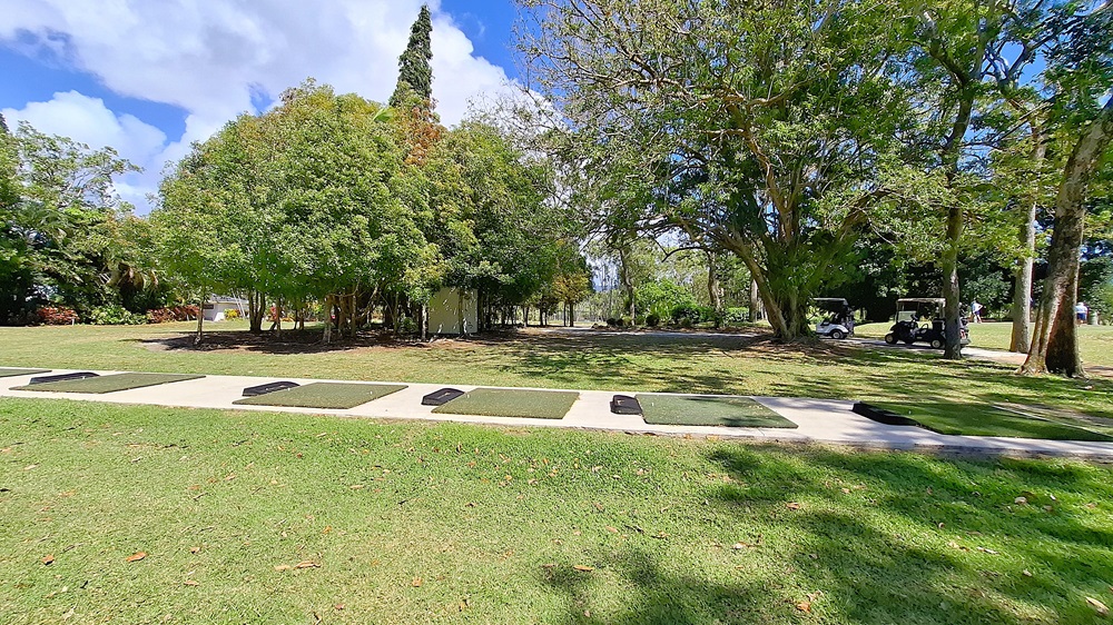 On the left, a well-maintained grassy area extends towards a line of trees, providing a natural boundary. In the centre, a gravel pathway leads into the distance, flanked by more greenery. On the right, a fenced area contains golf mats and a small practice area, with a few trees nearby. The ground is primarily gravel with patches of grass. On the left, a well-maintained grassy area extends towards a line of trees, providing a natural boundary. In the centre, a gravel pathway leads into the distance, flanked by more greenery. On the right, a fenced area contains golf mats and a small practice area, with a few trees nearby. The ground is primarily gravel with patches of grass.