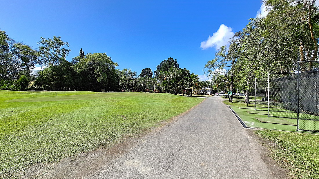 On the left, a large expanse of well-maintained grass stretches towards a line of trees, providing a natural backdrop. In the centre, a gravel pathway runs straight ahead, leading into the distance. On the right, a fenced area contains golf mats and a small practice space, with a few trees nearby. The ground is primarily gravel with patches of grass. On the left, a large expanse of well-maintained grass stretches towards a line of trees, providing a natural backdrop. In the centre, a gravel pathway runs straight ahead, leading into the distance. On the right, a fenced area contains golf mats and a small practice space, with a few trees nearby. The ground is primarily gravel with patches of grass.