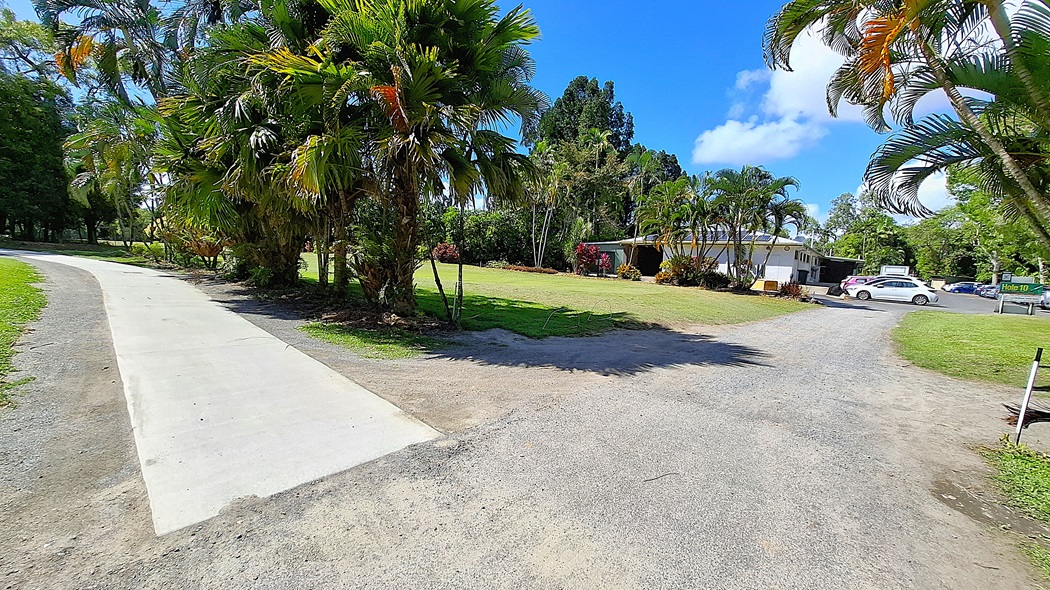 On the left, a concrete pathway curves gently, leading into a grassy area bordered by palm trees and shrubs. In the centre, a flat expanse of lawn is visible, with a small building partially obscured by greenery. On the right, a gravel road branches off, with a parked car and a sign indicating "Hole 10" nearby. The ground is a mix of concrete, gravel, and grass. On the left, a concrete pathway curves gently, leading into a grassy area bordered by palm trees and shrubs. In the centre, a flat expanse of lawn is visible, with a small building partially obscured by greenery. On the right, a gravel road branches off, with a parked car and a sign indicating "Hole 10" nearby. The ground is a mix of concrete, gravel, and grass.