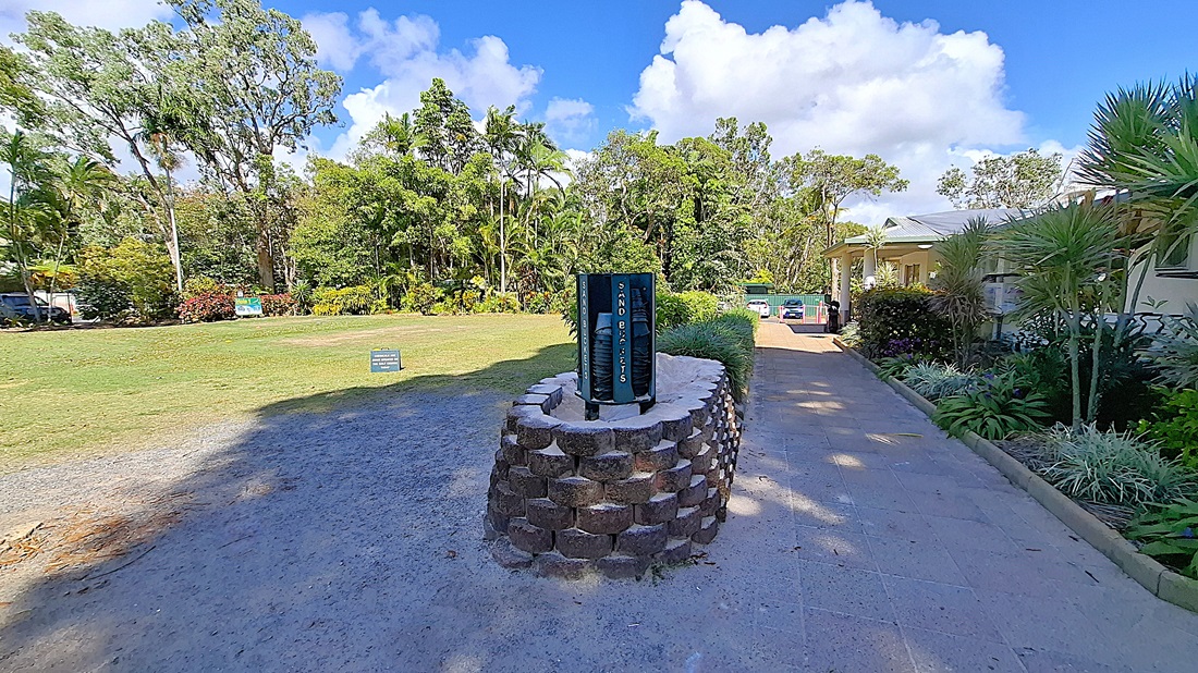 On the left, a grassy area is bordered by trees and shrubs, with a small sign indicating "Sand Tickets" positioned on the ground. The pathway is made of light-coloured tiles, leading towards a building on the right. In the centre, a circular stone feature stands with a vertical sign displaying information. On the right, the building has a light-coloured exterior and is surrounded by tropical plants. On the left, a grassy area is bordered by trees and shrubs, with a small sign indicating "Sand Tickets" positioned on the ground. The pathway is made of light-coloured tiles, leading towards a building on the right. In the centre, a circular stone feature stands with a vertical sign displaying information. On the right, the building has a light-coloured exterior and is surrounded by tropical plants.