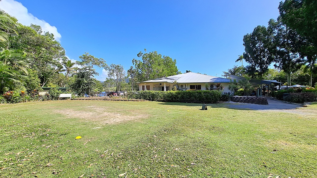 On the left, a grassy area is bordered by a variety of trees and shrubs, with a bench positioned for seating. The ground is mostly green, with some patches of bare earth. In the centre, a single-storey building features a light-coloured exterior and several air conditioning units mounted on the walls. On the right, a covered outdoor seating area is visible, surrounded by more greenery. The floor is a mix of grass and gravel. On the left, a grassy area is bordered by a variety of trees and shrubs, with a bench positioned for seating. The ground is mostly green, with some patches of bare earth. In the centre, a single-storey building features a light-coloured exterior and several air conditioning units mounted on the walls. On the right, a covered outdoor seating area is visible, surrounded by more greenery. The floor is a mix of grass and gravel.