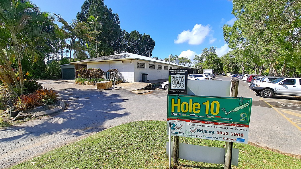 On the left, a building with a light-coloured exterior features several windows and a door, with a small area of landscaping including shrubs and a palm tree. A bin is positioned next to the building. In the centre, a sign reading "Hole 10" is prominently displayed, indicating the golf course hole details. On the right, a parking area is visible with multiple vehicles parked in a gravel lot. The ground is a mix of gravel and grass. On the left, a building with a light-coloured exterior features several windows and a door, with a small area of landscaping including shrubs and a palm tree. A bin is positioned next to the building. In the centre, a sign reading "Hole 10" is prominently displayed, indicating the golf course hole details. On the right, a parking area is visible with multiple vehicles parked in a gravel lot. The ground is a mix of gravel and grass.