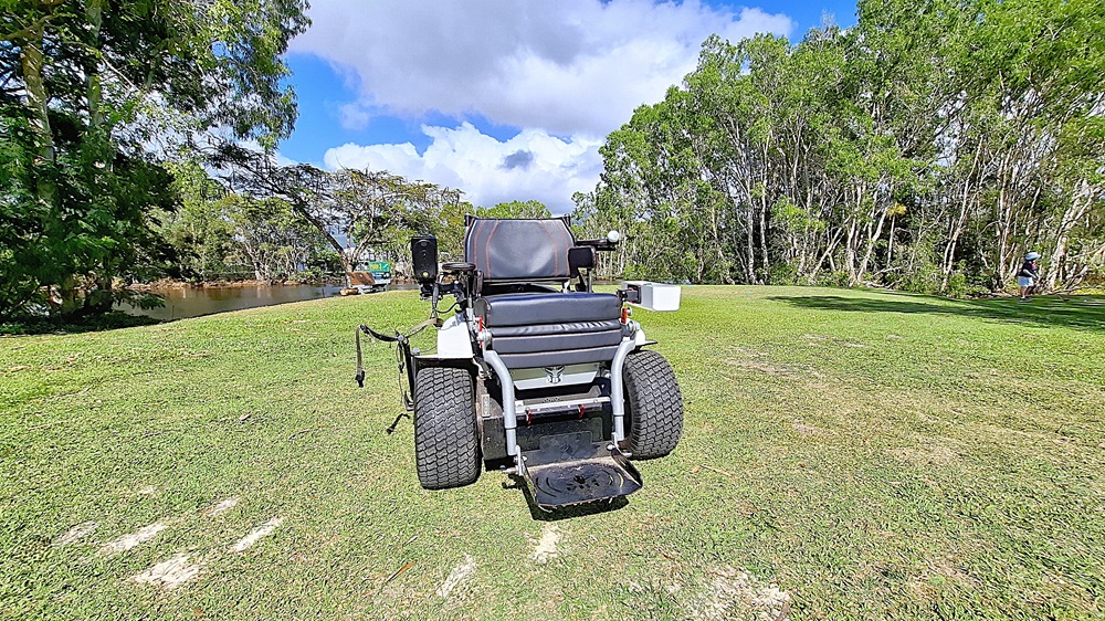 On the left, a patch of grass is visible, bordered by trees that provide shade. A small body of water can be seen in the background, reflecting the greenery. In the centre, a golf cart is parked, featuring a black seat and large wheels, with controls visible on the sides. On the right, a person is walking in the distance, partially obscured by trees. The ground is a well-maintained lawn. On the left, a patch of grass is visible, bordered by trees that provide shade. A small body of water can be seen in the background, reflecting the greenery. In the centre, a golf cart is parked, featuring a black seat and large wheels, with controls visible on the sides. On the right, a person is walking in the distance, partially obscured by trees. The ground is a well-maintained lawn.