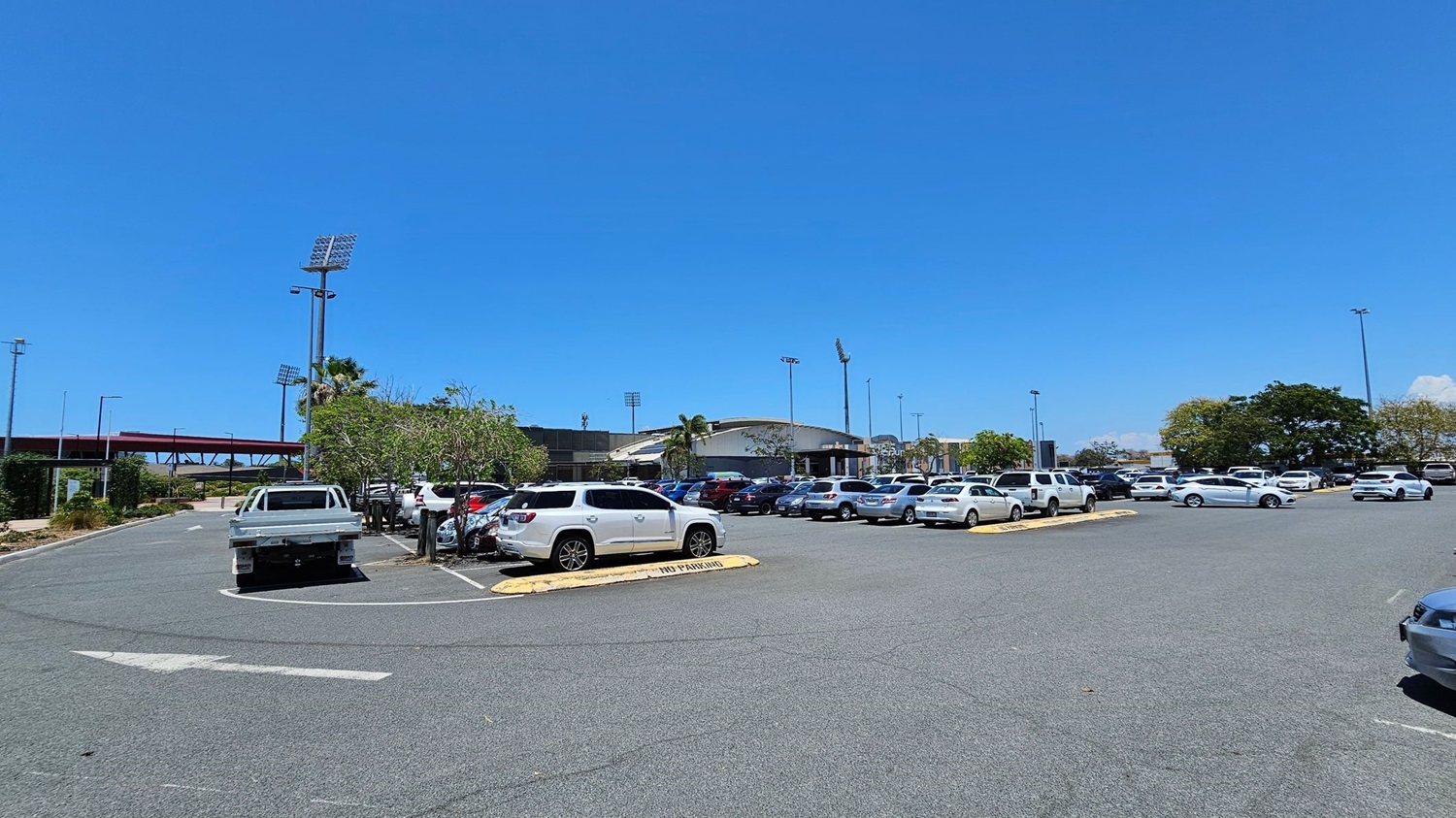 A large car park with numerous vehicles, including white and silver cars, is in the foreground. In the background, there is a stadium with tall floodlights and a curved roof. Trees and small shrubs are scattered around the parking area. A red pedestrian bridge is visible on the left side, and the ground is paved with asphalt.