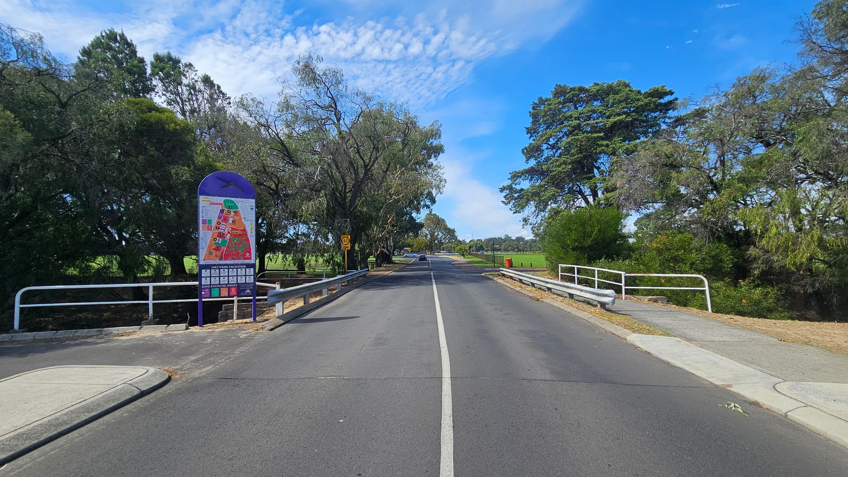 A straight road with a white centre line runs through the middle, flanked by metal railings on both sides. On the left, there's a colourful map sign with trees behind it. On the right, more trees and a red bin are visible. The road leads into a park-like area with more greenery and a few distant cars. The sky is blue with scattered clouds. A straight road with a white centre line runs through the middle, flanked by metal railings on both sides. On the left, there's a colourful map sign with trees behind it. On the right, more trees and a red bin are visible. The road leads into a park-like area with more greenery and a few distant cars. The sky is blue with scattered clouds.
