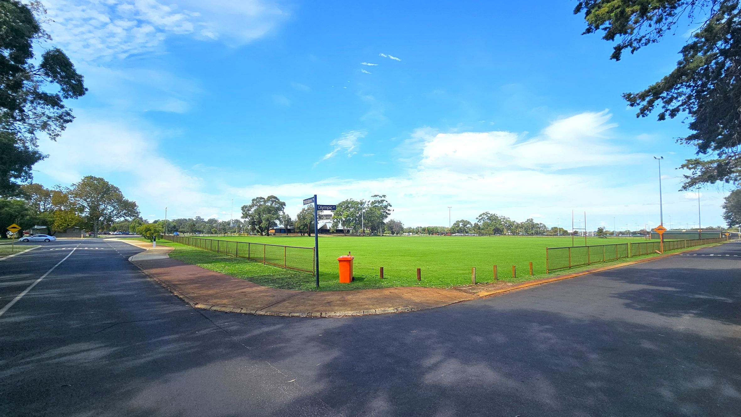 A wide, open grassy field is in the centre, surrounded by a low metal fence. An orange bin is near the fence. To the left, a paved road curves with a few parked cars visible. Trees line the background, providing a natural border. The sky is clear with a few clouds. The foreground shows a paved area with shadows from nearby trees. A wide, open grassy field is in the centre, surrounded by a low metal fence. An orange bin is near the fence. To the left, a paved road curves with a few parked cars visible. Trees line the background, providing a natural border. The sky is clear with a few clouds. The foreground shows a paved area with shadows from nearby trees.