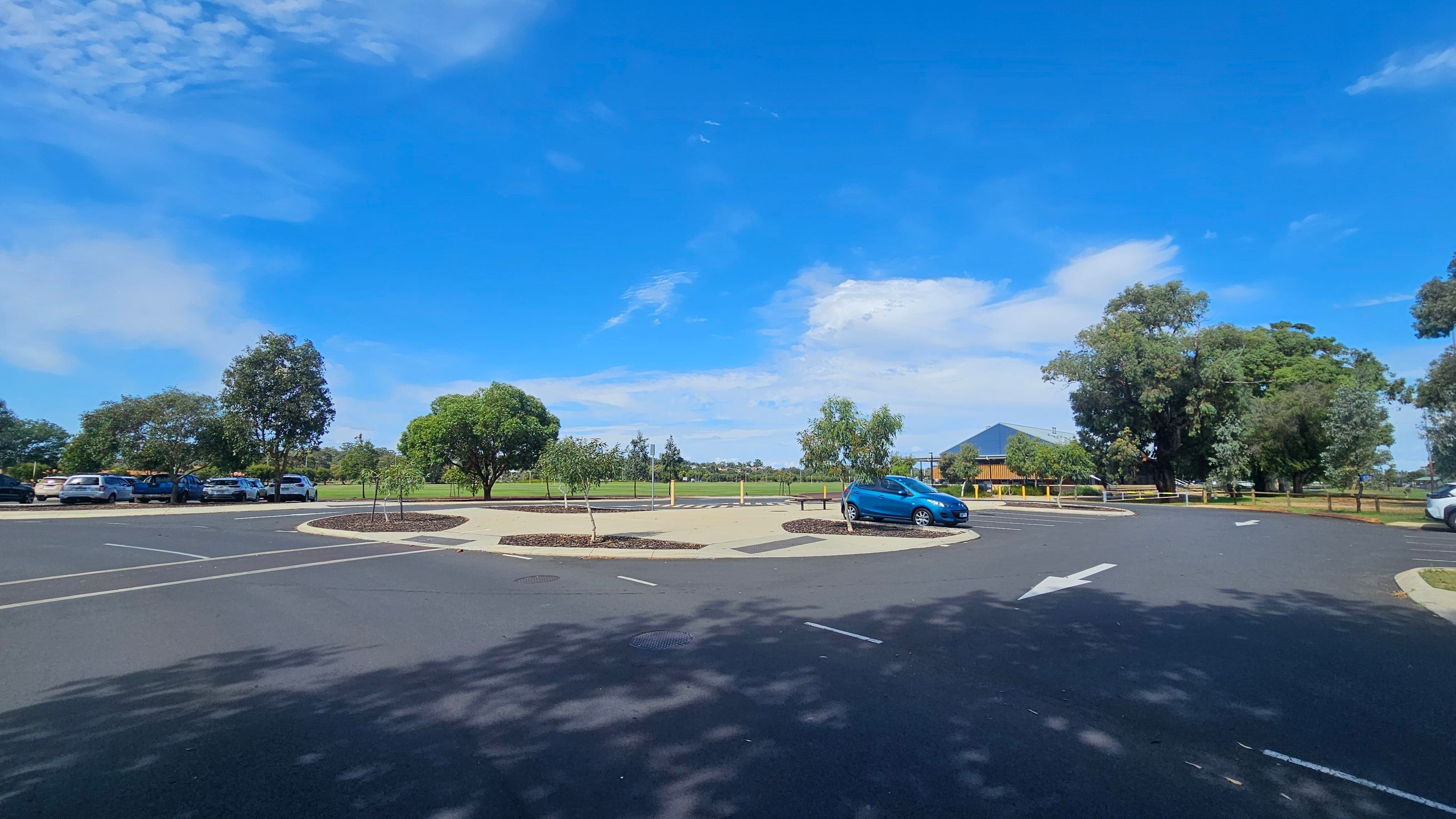 A car park with a roundabout in the centre, featuring a few small trees and a blue car parked on the right. On the left, several cars are parked along the edge, with trees and greenery in the background. A building with a dark roof is visible in the distance on the right. The surface is paved with marked parking spaces and directional arrows. A car park with a roundabout in the centre, featuring a few small trees and a blue car parked on the right. On the left, several cars are parked along the edge, with trees and greenery in the background. A building with a dark roof is visible in the distance on the right. The surface is paved with marked parking spaces and directional arrows.