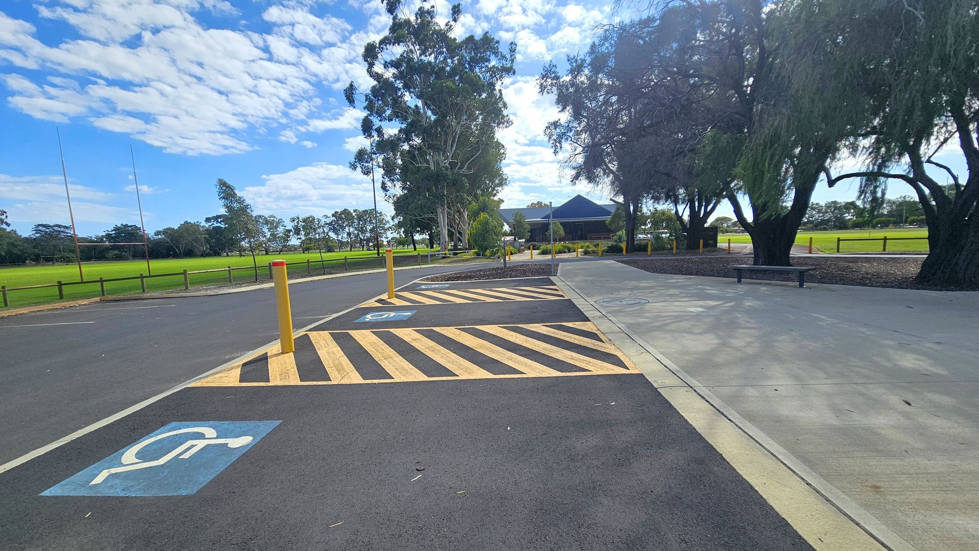 A paved area with a marked disabled parking space on the left, featuring a blue symbol. Yellow and black striped lines are adjacent to the parking space. A yellow bollard stands nearby. On the right, a concrete path runs alongside trees with dense foliage. In the background, a grassy field with rugby posts is visible, along with a building partially obscured by trees. A bench is positioned near the path. A paved area with a marked disabled parking space on the left, featuring a blue symbol. Yellow and black striped lines are adjacent to the parking space. A yellow bollard stands nearby. On the right, a concrete path runs alongside trees with dense foliage. In the background, a grassy field with rugby posts is visible, along with a building partially obscured by trees. A bench is positioned near the path.