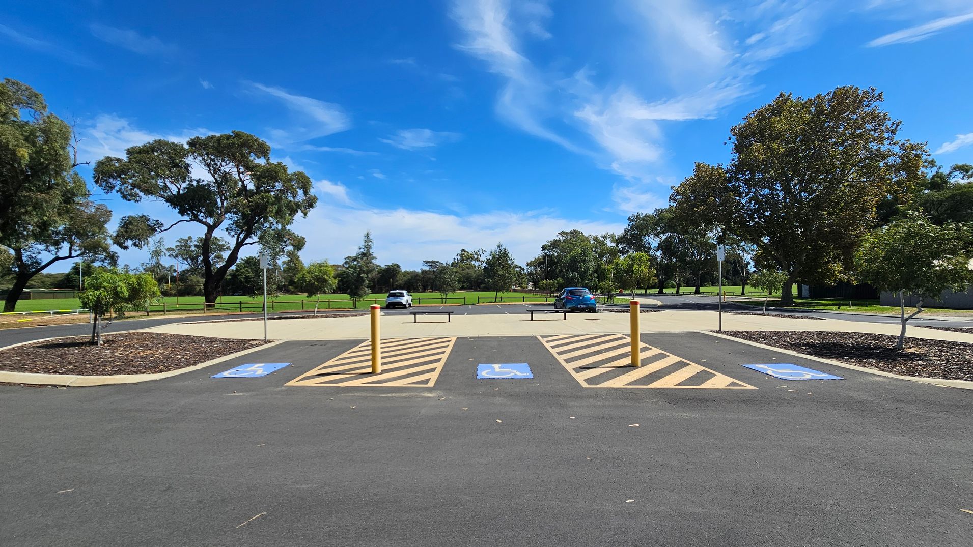 A car park with designated disabled parking spaces marked by blue signs on the asphalt. Yellow bollards separate the spaces. In the background, there are two cars parked, one white and one blue. Surrounding the car park are green grassy areas and several large trees. The sky is clear with some wispy clouds. A car park with designated disabled parking spaces marked by blue signs on the asphalt. Yellow bollards separate the spaces. In the background, there are two cars parked, one white and one blue. Surrounding the car park are green grassy areas and several large trees. The sky is clear with some wispy clouds.
