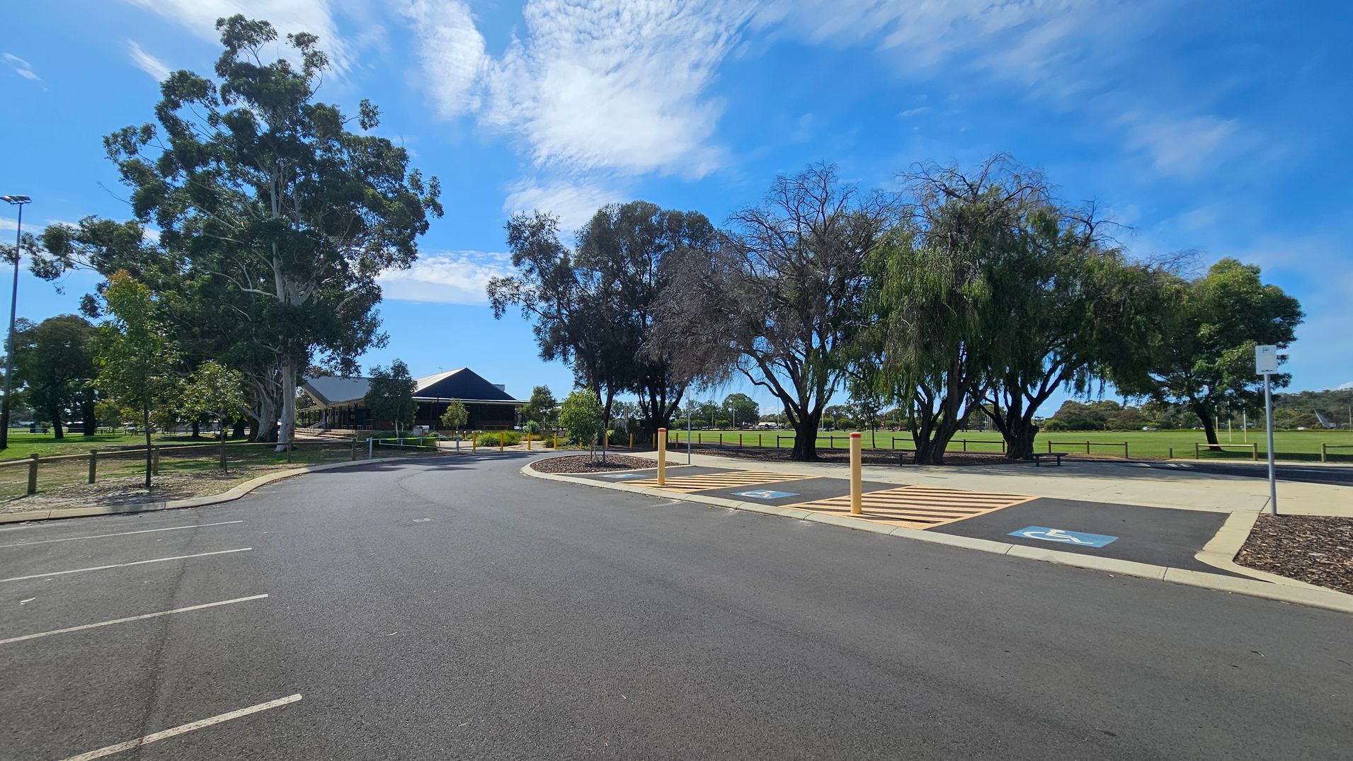 A paved parking area with marked spaces, including a designated disabled parking spot on the right. A row of tall trees lines the background, with a building partially visible to the left. The area is bordered by a grassy field and wooden fencing. The sky is clear with some clouds. A paved parking area with marked spaces, including a designated disabled parking spot on the right. A row of tall trees lines the background, with a building partially visible to the left. The area is bordered by a grassy field and wooden fencing. The sky is clear with some clouds.
