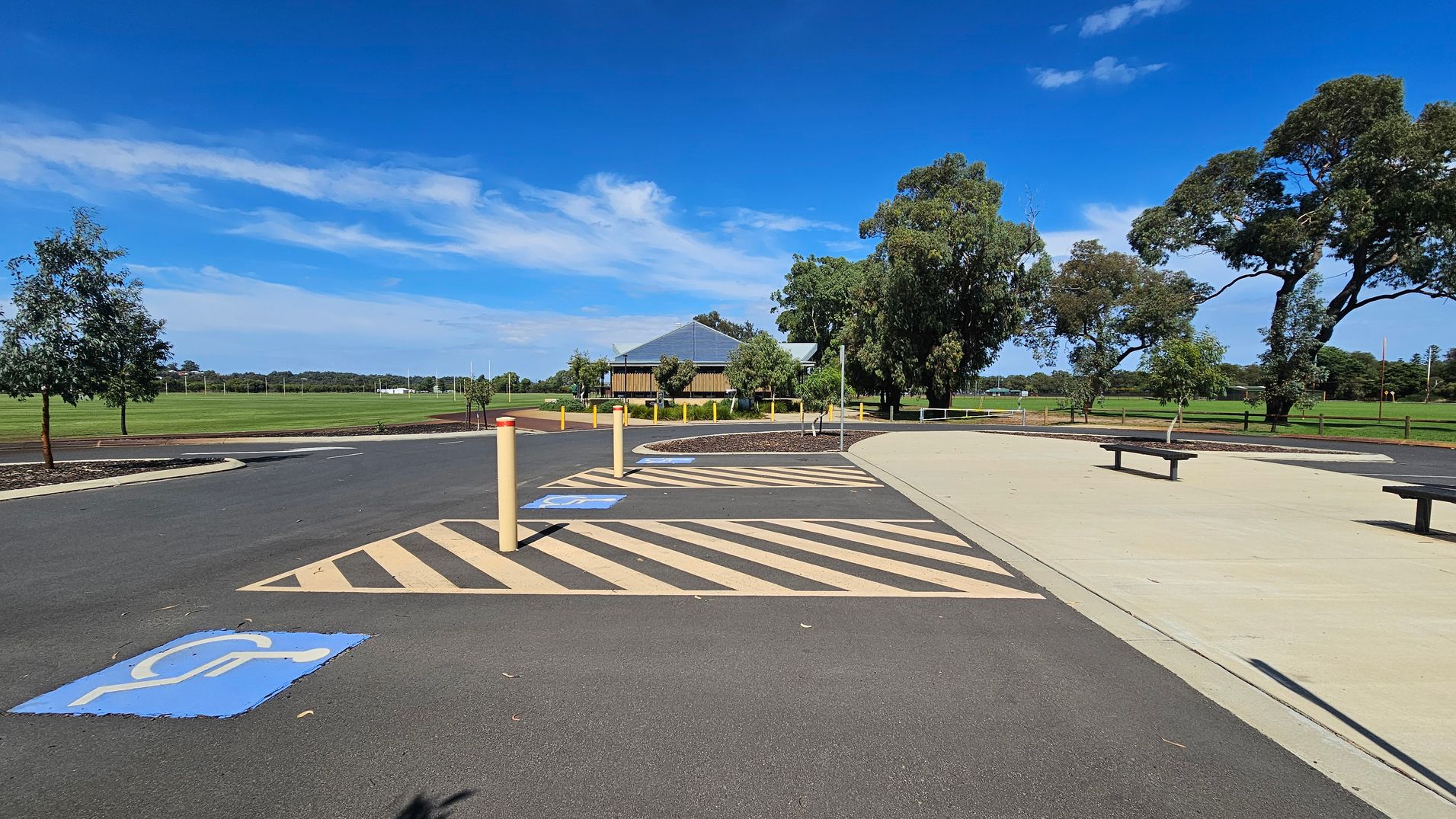 A paved parking area with marked disabled parking spaces and yellow-striped zones. In the centre, a small building with a sloped roof is surrounded by trees. To the right, a concrete path with benches runs parallel to the parking area. The background features a large grassy field with more trees and a fence. The sky is clear with some clouds. A paved parking area with marked disabled parking spaces and yellow-striped zones. In the centre, a small building with a sloped roof is surrounded by trees. To the right, a concrete path with benches runs parallel to the parking area. The background features a large grassy field with more trees and a fence. The sky is clear with some clouds.