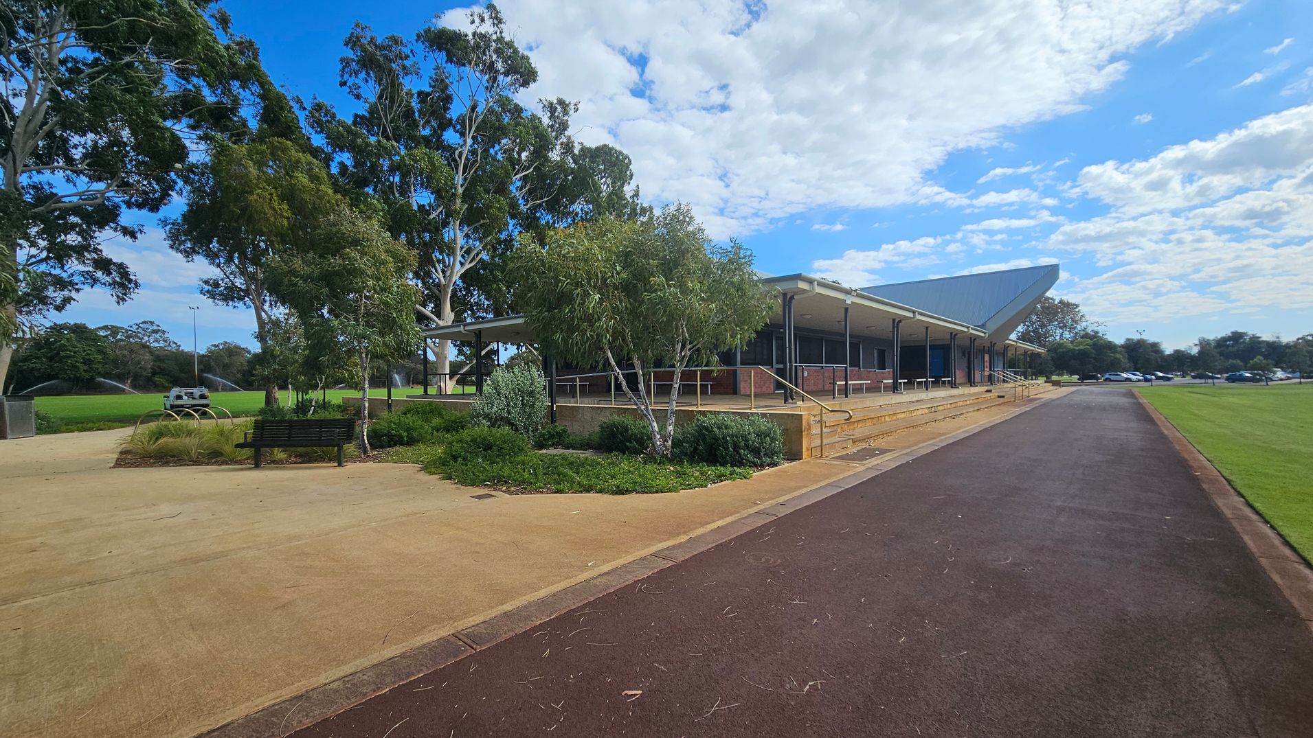 A paved path runs through the foreground, leading to a building on the right with a sloped roof and a series of steps. The building is surrounded by a landscaped area with shrubs and small trees. On the left, there is a bench and more greenery, with large trees in the background. A grassy field with sprinklers is visible to the left, and several parked cars can be seen in the distance on the right. A paved path runs through the foreground, leading to a building on the right with a sloped roof and a series of steps. The building is surrounded by a landscaped area with shrubs and small trees. On the left, there is a bench and more greenery, with large trees in the background. A grassy field with sprinklers is visible to the left, and several parked cars can be seen in the distance on the right.