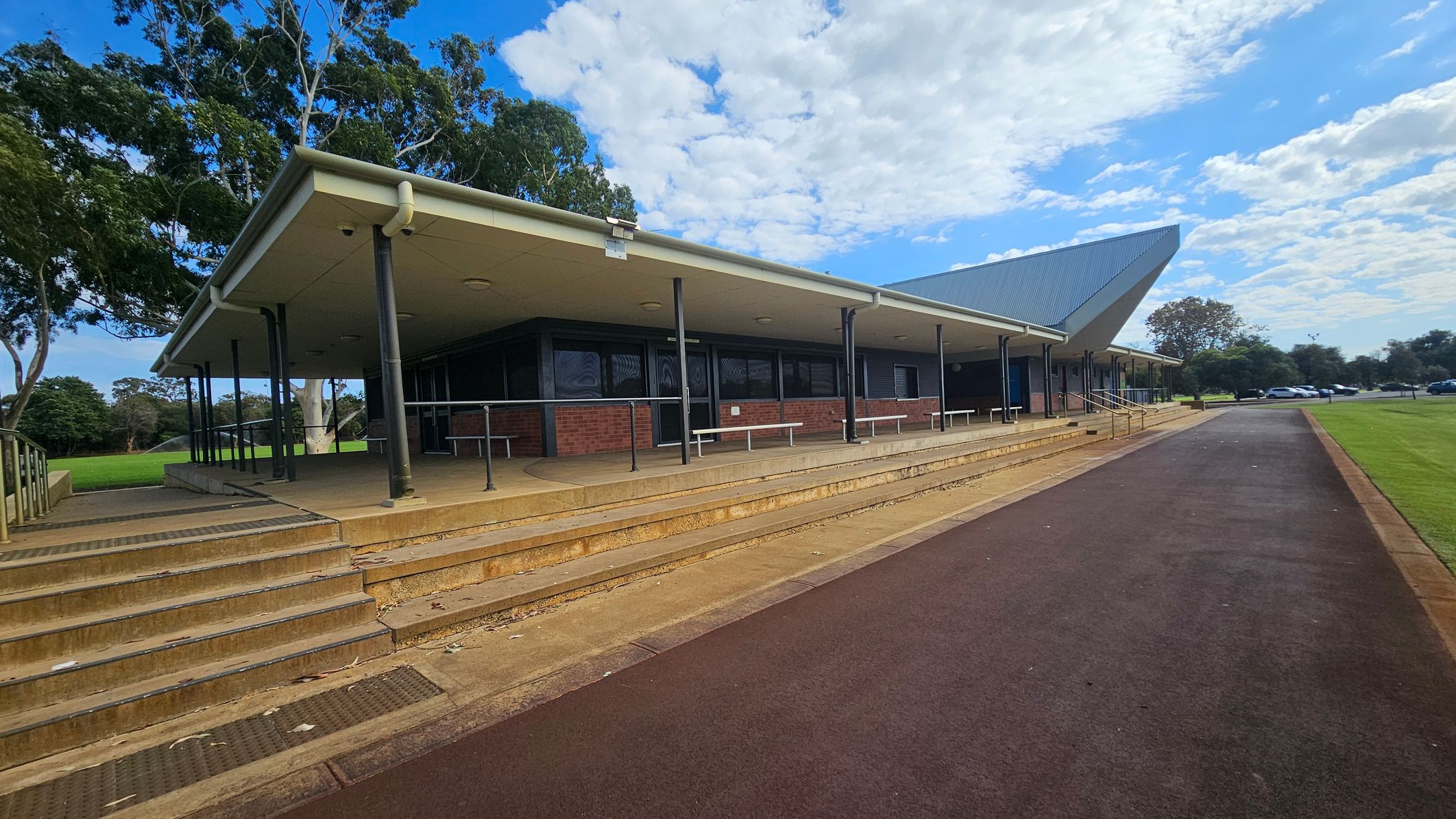 A modern building with a sloped roof and a covered walkway supported by black poles. The structure is made of brick with large windows. In the foreground, there are steps leading up to the entrance and a ramp with railings. The ground is paved with a reddish-brown path running alongside a grassy area. Trees are visible in the background, and several cars are parked in the distance. The sky is partly cloudy. A modern building with a sloped roof and a covered walkway supported by black poles. The structure is made of brick with large windows. In the foreground, there are steps leading up to the entrance and a ramp with railings. The ground is paved with a reddish-brown path running alongside a grassy area. Trees are visible in the background, and several cars are parked in the distance. The sky is partly cloudy.