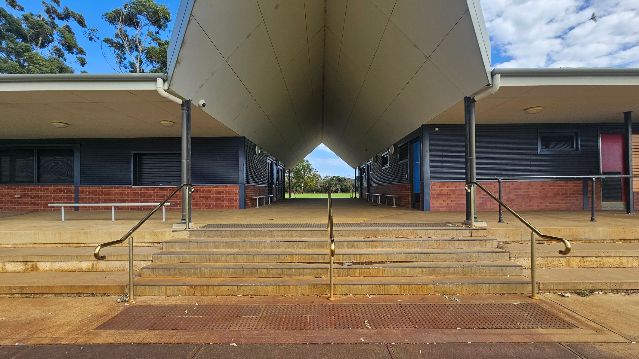 A symmetrical view of a building entrance with a wide, covered walkway leading to an open field in the background. The structure has a sloped roof supported by black columns. The walls are a combination of dark panels and red brick. Steps with metal railings lead up to the entrance. Benches are positioned along the walls on both sides. The ceiling is flat and light-coloured, with circular lights. Trees are visible in the distance beyond the walkway. A symmetrical view of a building entrance with a wide, covered walkway leading to an open field in the background. The structure has a sloped roof supported by black columns. The walls are a combination of dark panels and red brick. Steps with metal railings lead up to the entrance. Benches are positioned along the walls on both sides. The ceiling is flat and light-coloured, with circular lights. Trees are visible in the distance beyond the walkway.
