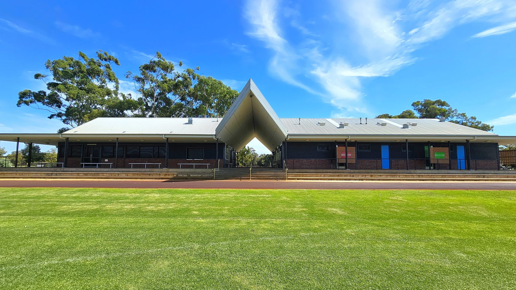 A building with a distinctive V-shaped roof structure in the centre, flanked by two sections with grey roofs. The building has dark walls with blue and green doors. In the foreground, there is a well-maintained grassy area. Trees are visible in the background on both sides. A building with a distinctive V-shaped roof structure in the centre, flanked by two sections with grey roofs. The building has dark walls with blue and green doors. In the foreground, there is a well-maintained grassy area. Trees are visible in the background on both sides.