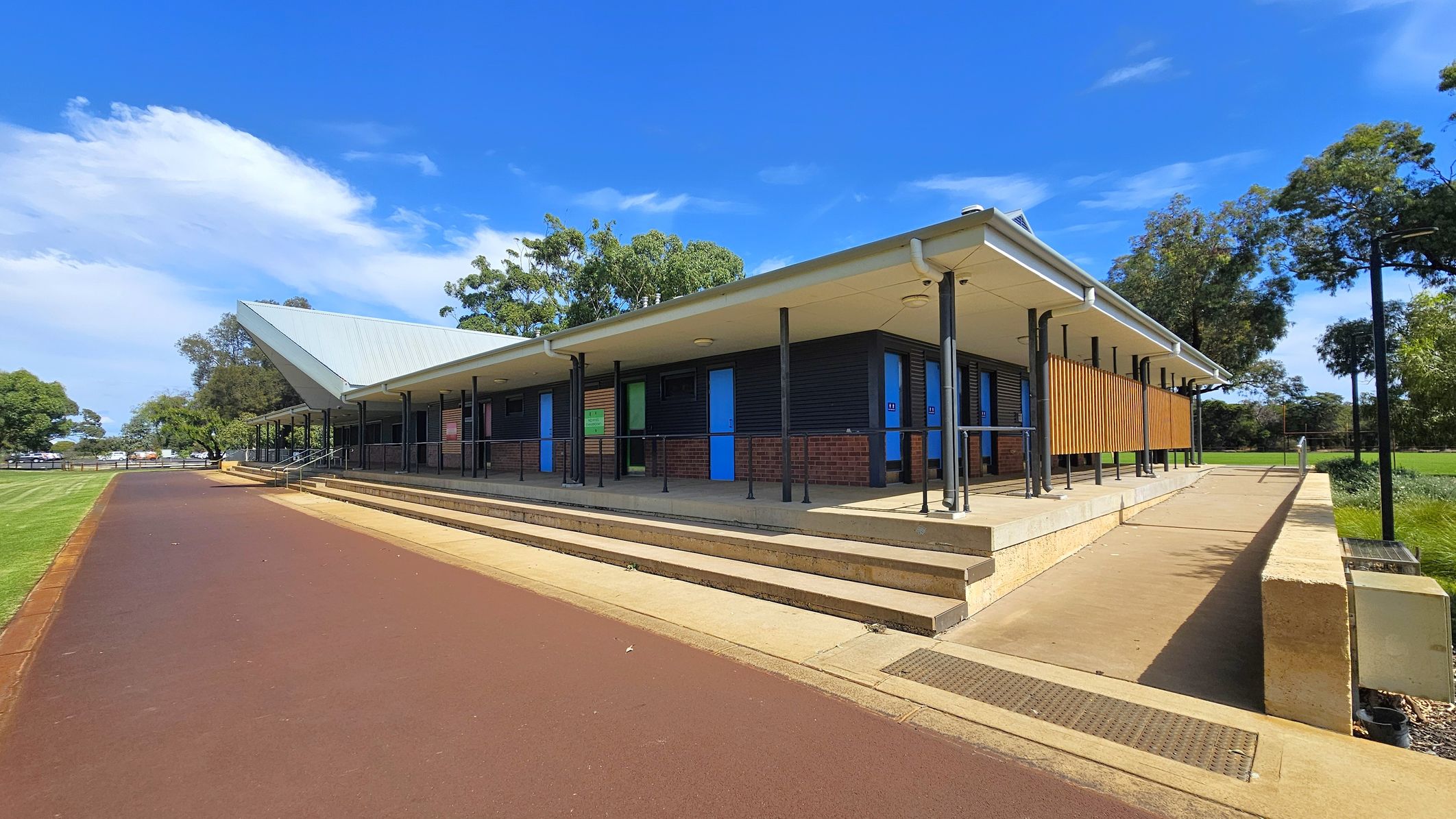 A modern building with a sloped metal roof and a covered walkway supported by black poles. The structure features a combination of brick and dark panelling, with bright blue doors. A series of steps and a ramp lead up to the entrance. On the left, a wide, reddish-brown path runs alongside a grassy area. Trees are visible in the background, providing a natural setting around the building. A modern building with a sloped metal roof and a covered walkway supported by black poles. The structure features a combination of brick and dark panelling, with bright blue doors. A series of steps and a ramp lead up to the entrance. On the left, a wide, reddish-brown path runs alongside a grassy area. Trees are visible in the background, providing a natural setting around the building.