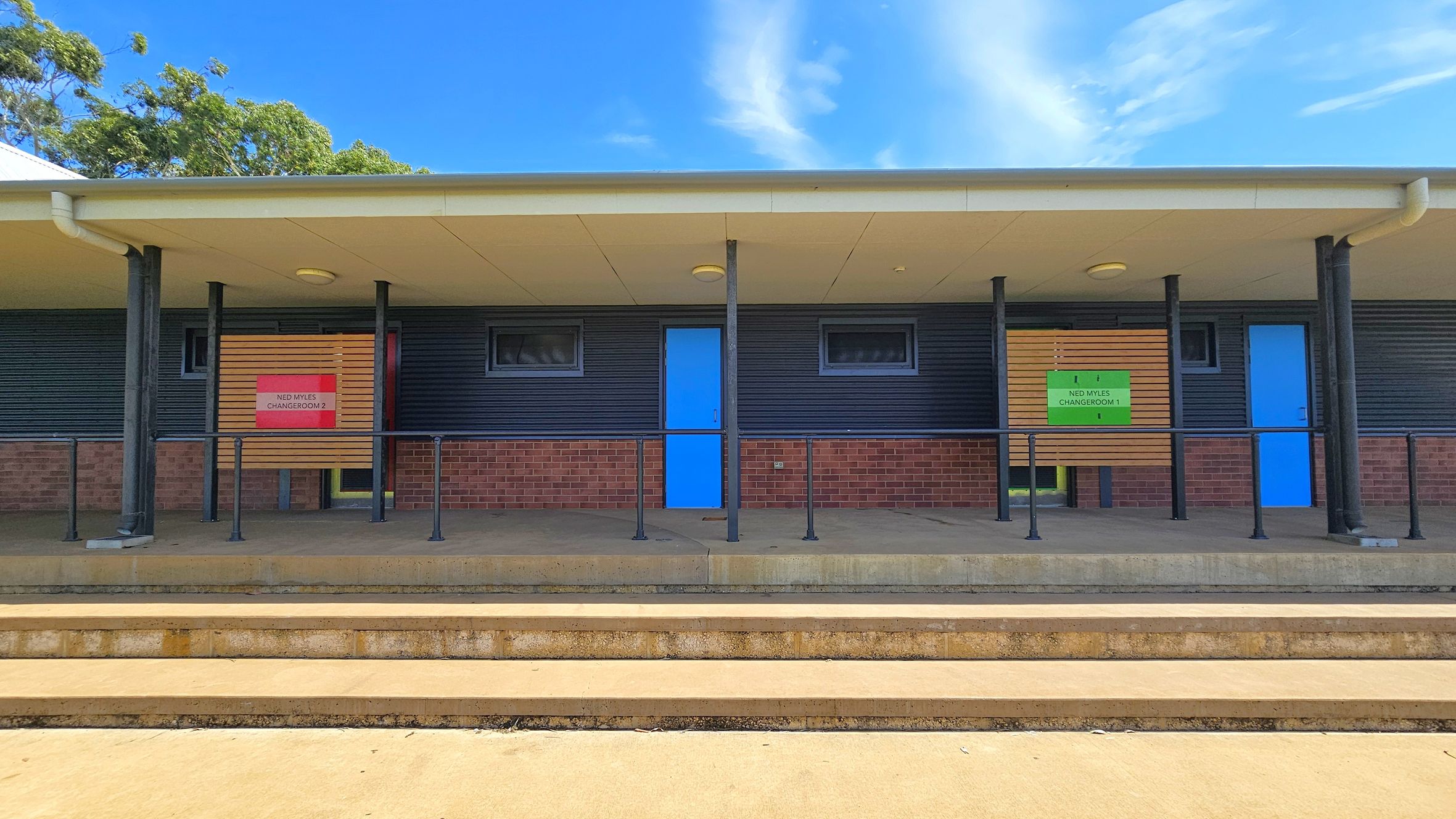 A building with a brick lower half and dark corrugated upper half. Two blue doors are centred between small windows. Signs above the doors read "Ned Myles Changeroom 2" on the left and "Ned Myles Changeroom 1" on the right. The roof is supported by black poles. The foreground features concrete steps leading up to the building. Trees are visible in the background. A building with a brick lower half and dark corrugated upper half. Two blue doors are centred between small windows. Signs above the doors read "Ned Myles Changeroom 2" on the left and "Ned Myles Changeroom 1" on the right. The roof is supported by black poles. The foreground features concrete steps leading up to the building. Trees are visible in the background.