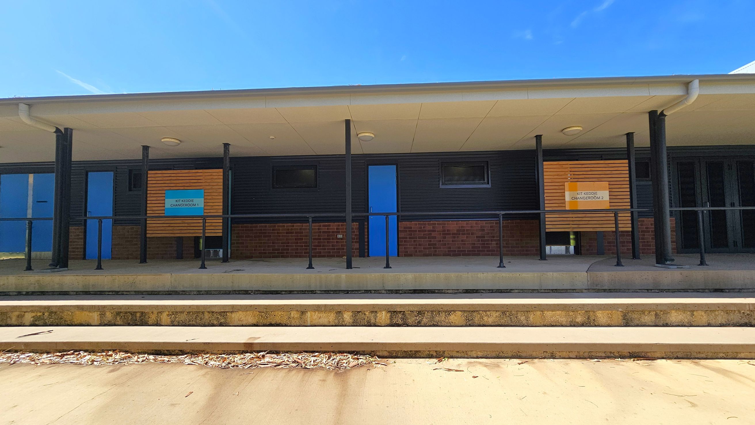 A building with a covered walkway supported by black poles. The facade features dark cladding and brickwork. Two blue doors are visible, each flanked by small rectangular windows. Signs above the doors read "Kit Keddie Changeroom 1" and "Kit Keddie Changeroom 2". The foreground has concrete steps leading up to the building, with some dried leaves scattered on the ground. The ceiling of the walkway is light-coloured with circular lights. A building with a covered walkway supported by black poles. The facade features dark cladding and brickwork. Two blue doors are visible, each flanked by small rectangular windows. Signs above the doors read "Kit Keddie Changeroom 1" and "Kit Keddie Changeroom 2". The foreground has concrete steps leading up to the building, with some dried leaves scattered on the ground. The ceiling of the walkway is light-coloured with circular lights.
