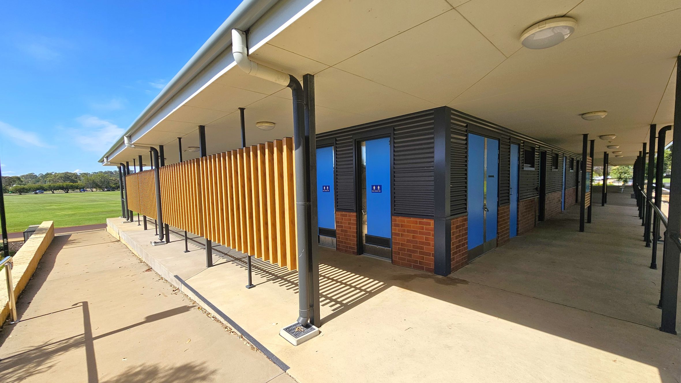 A building with a sloped roof and a series of blue doors on the right, likely public toilets, with signage indicating male and female facilities. The structure features a combination of brick and corrugated metal walls. Wooden slats run vertically along part of the exterior. The foreground has a concrete path with a metal railing, leading to a grassy field in the background. The ceiling has circular lights, and the area is supported by black metal poles. A building with a sloped roof and a series of blue doors on the right, likely public toilets, with signage indicating male and female facilities. The structure features a combination of brick and corrugated metal walls. Wooden slats run vertically along part of the exterior. The foreground has a concrete path with a metal railing, leading to a grassy field in the background. The ceiling has circular lights, and the area is supported by black metal poles.