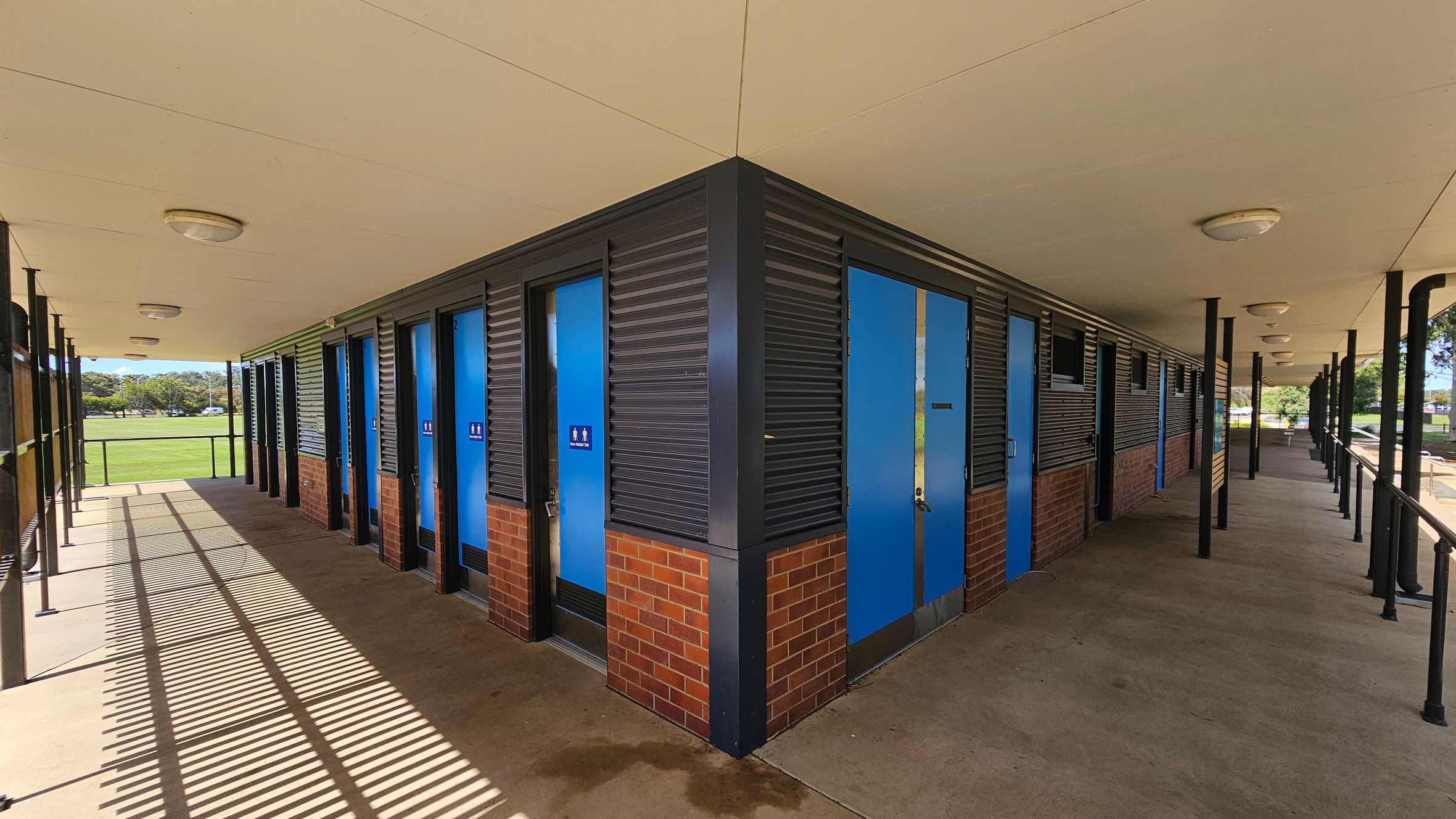 A building with a corner structure featuring blue doors and brick walls. The left side has a row of doors with signs, under a covered walkway with a concrete floor. The ceiling is white with round lights. The right side also has doors and windows, with black metal railings along the walkway. In the background, there is a grassy field and trees. Shadows from the railing create patterns on the floor. A building with a corner structure featuring blue doors and brick walls. The left side has a row of doors with signs, under a covered walkway with a concrete floor. The ceiling is white with round lights. The right side also has doors and windows, with black metal railings along the walkway. In the background, there is a grassy field and trees. Shadows from the railing create patterns on the floor.