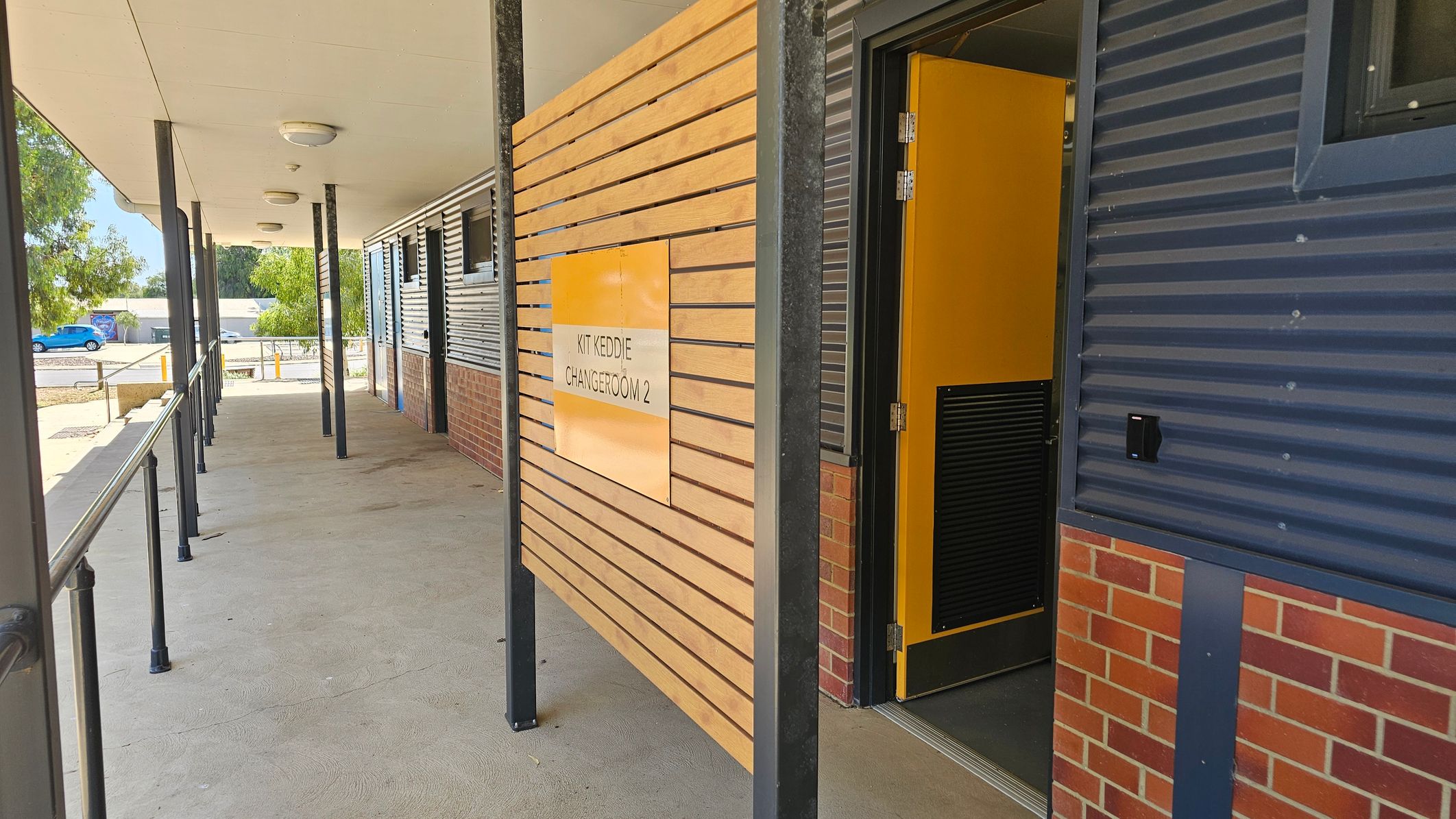 A corridor with a covered walkway featuring a series of black metal poles supporting the ceiling. On the right, a wooden panel with horizontal slats displays a sign for "Kit Keddie Changeroom 2" next to an open yellow door. The building has a combination of corrugated metal and brick walls. The floor is concrete with a greenish tint. In the background, there are trees and a blue car parked near some bollards. A corridor with a covered walkway featuring a series of black metal poles supporting the ceiling. On the right, a wooden panel with horizontal slats displays a sign for "Kit Keddie Changeroom 2" next to an open yellow door. The building has a combination of corrugated metal and brick walls. The floor is concrete with a greenish tint. In the background, there are trees and a blue car parked near some bollards.