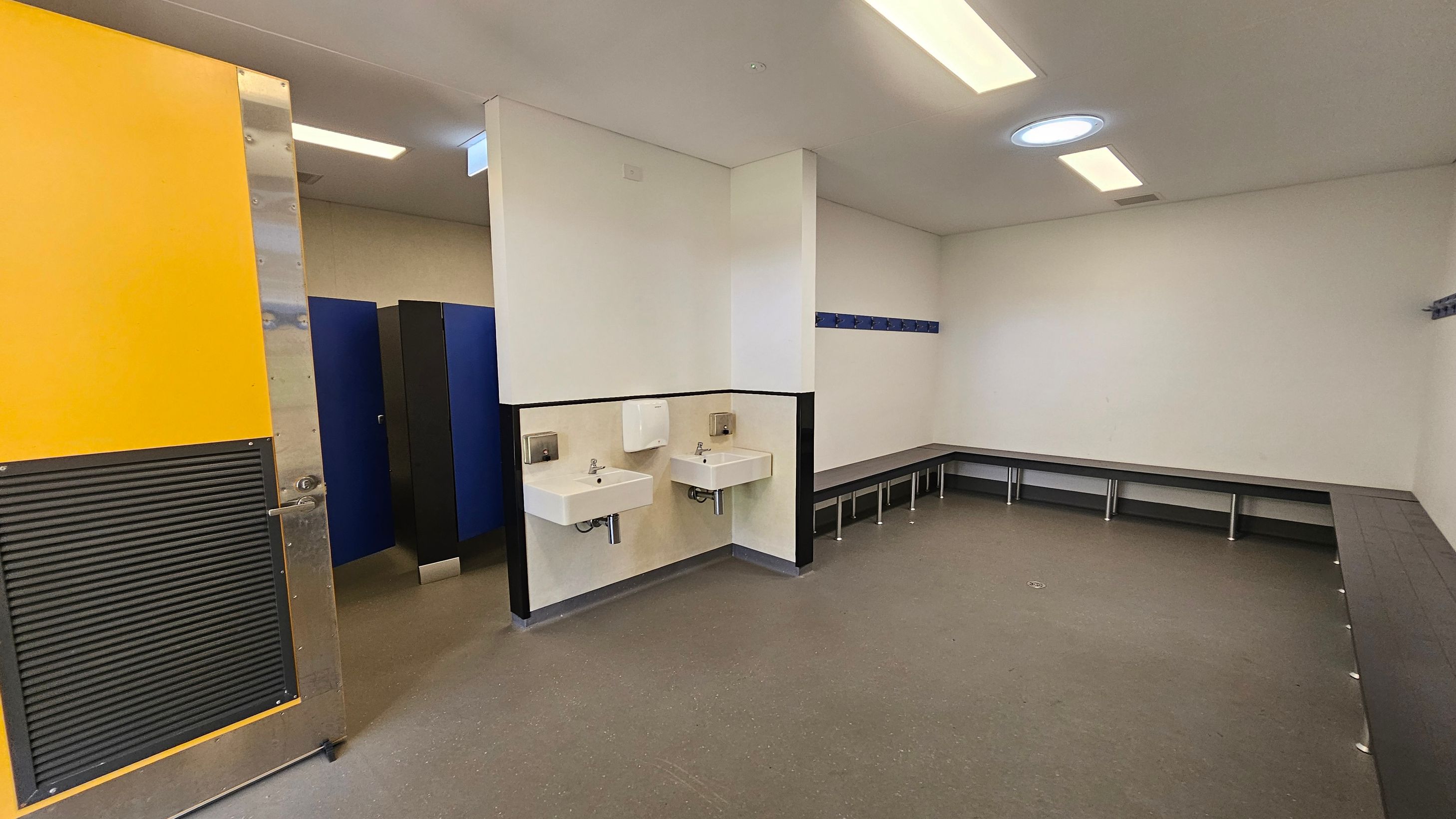 A changing room with a yellow door on the left, partially open, revealing blue lockers. In the centre, two white sinks are mounted on the wall with a paper towel dispenser above. On the right, a long bench runs along the walls, with hooks above for hanging items. The floor is grey, and the ceiling has rectangular lights and a circular skylight. A changing room with a yellow door on the left, partially open, revealing blue lockers. In the centre, two white sinks are mounted on the wall with a paper towel dispenser above. On the right, a long bench runs along the walls, with hooks above for hanging items. The floor is grey, and the ceiling has rectangular lights and a circular skylight.