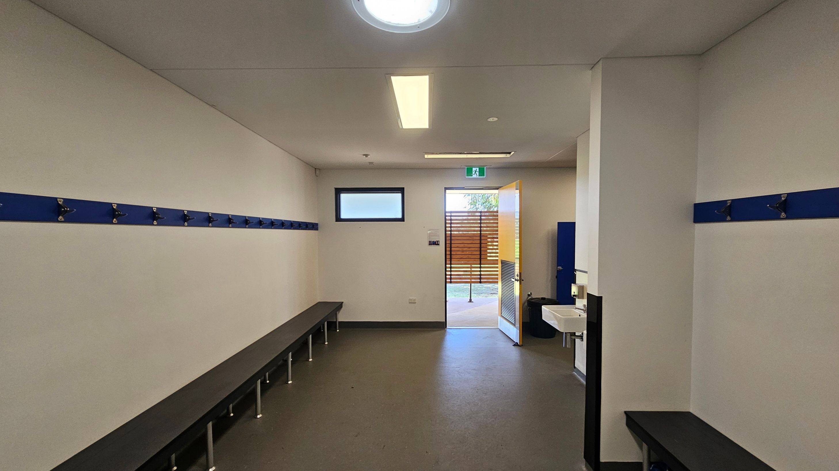A changing room with white walls and a grey floor. On the left and right walls, there are blue hooks for hanging items, with long benches beneath them. In the middle, an open door leads outside, allowing natural light to enter. A small window is above the door. On the right, a white sink is mounted on the wall. The ceiling has a circular light fixture and a rectangular fluorescent light. A changing room with white walls and a grey floor. On the left and right walls, there are blue hooks for hanging items, with long benches beneath them. In the middle, an open door leads outside, allowing natural light to enter. A small window is above the door. On the right, a white sink is mounted on the wall. The ceiling has a circular light fixture and a rectangular fluorescent light.