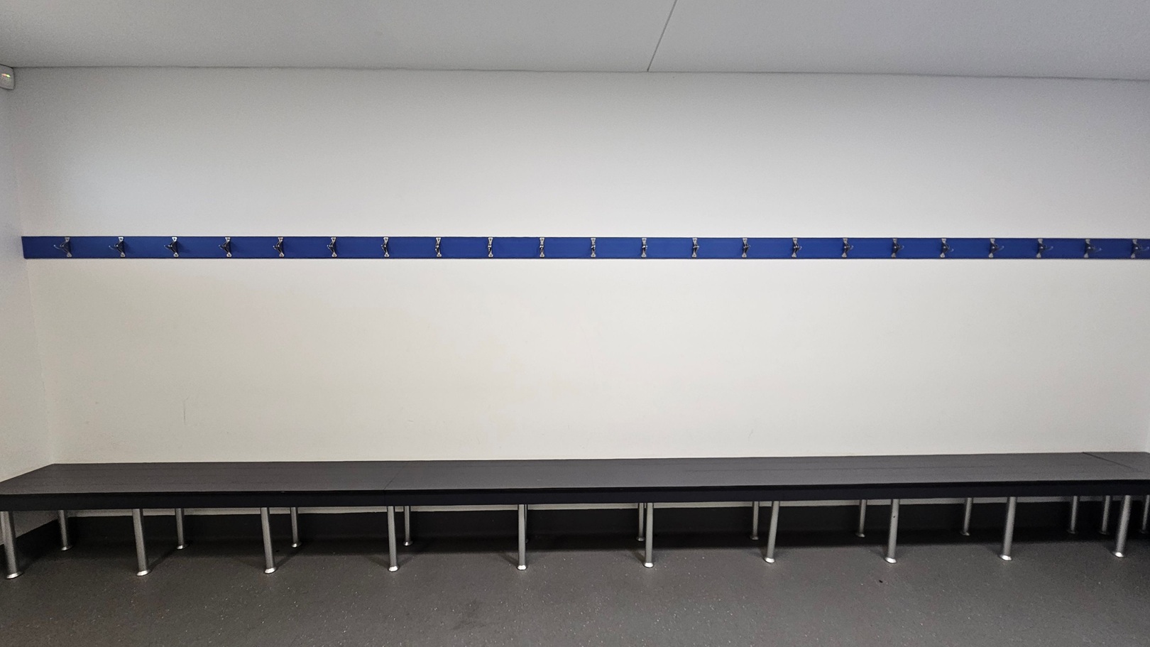 A room with a long, dark bench along the wall, supported by metal legs. Above the bench, a blue strip with multiple evenly spaced hooks is mounted on the white wall. The floor is grey and speckled, and the ceiling is plain white. The room appears to be a changing or locker room. A room with a long, dark bench along the wall, supported by metal legs. Above the bench, a blue strip with multiple evenly spaced hooks is mounted on the white wall. The floor is grey and speckled, and the ceiling is plain white. The room appears to be a changing or locker room.