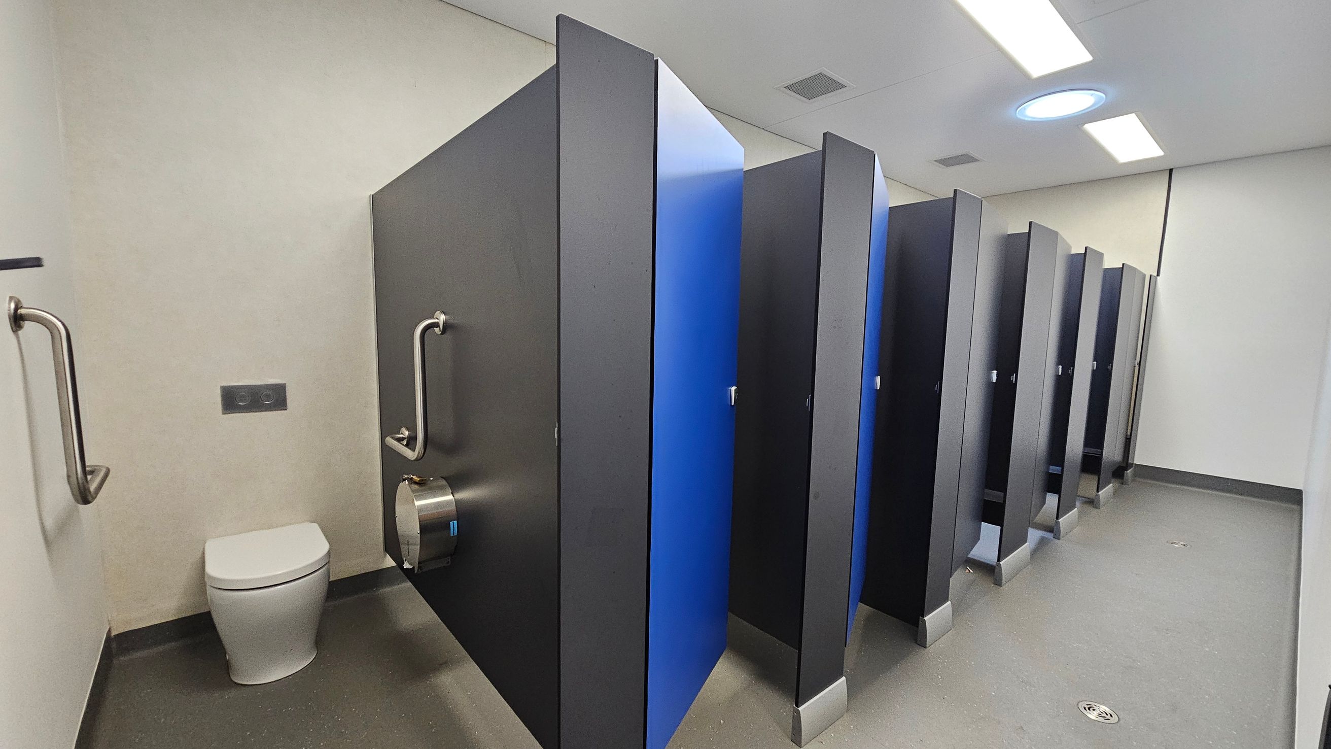 A public restroom with a row of toilet cubicles on the right, featuring dark grey partitions and blue doors. On the left, there is a single toilet with grab bars on either side and a toilet paper dispenser attached to the partition. The floor is grey with a drain visible, and the ceiling is white with rectangular and circular lights. A public restroom with a row of toilet cubicles on the right, featuring dark grey partitions and blue doors. On the left, there is a single toilet with grab bars on either side and a toilet paper dispenser attached to the partition. The floor is grey with a drain visible, and the ceiling is white with rectangular and circular lights.