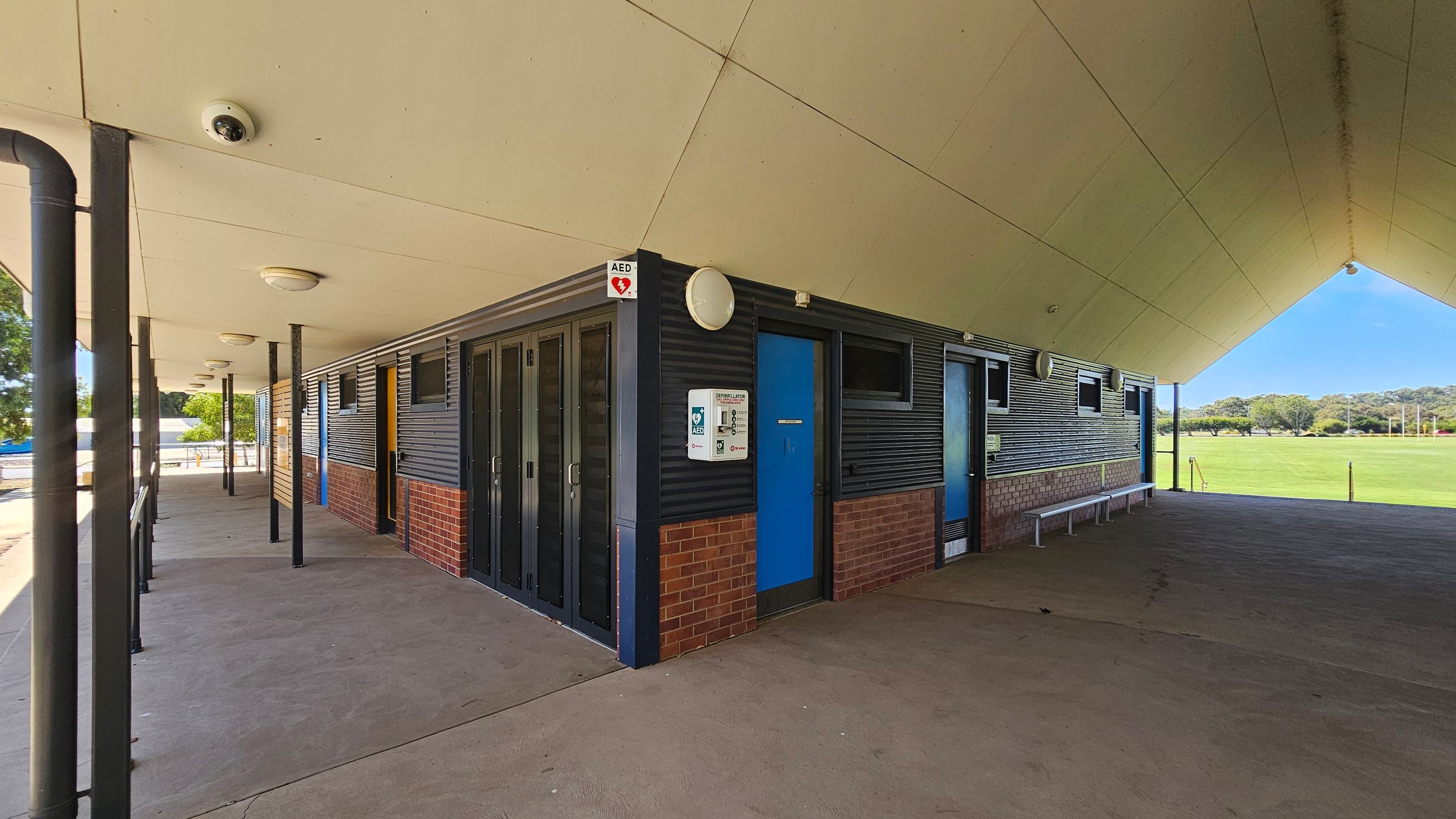 A building with a sloped roof and a covered walkway. The exterior walls are a combination of dark panels and red brick. A blue door is visible on the right, with a sign for an AED (Automated External Defibrillator) mounted nearby. Several windows are along the wall. The ceiling is light-coloured with circular lights. The floor is concrete, and there are metal benches along the wall. In the background, there is a grassy field with trees. A building with a sloped roof and a covered walkway. The exterior walls are a combination of dark panels and red brick. A blue door is visible on the right, with a sign for an AED (Automated External Defibrillator) mounted nearby. Several windows are along the wall. The ceiling is light-coloured with circular lights. The floor is concrete, and there are metal benches along the wall. In the background, there is a grassy field with trees.